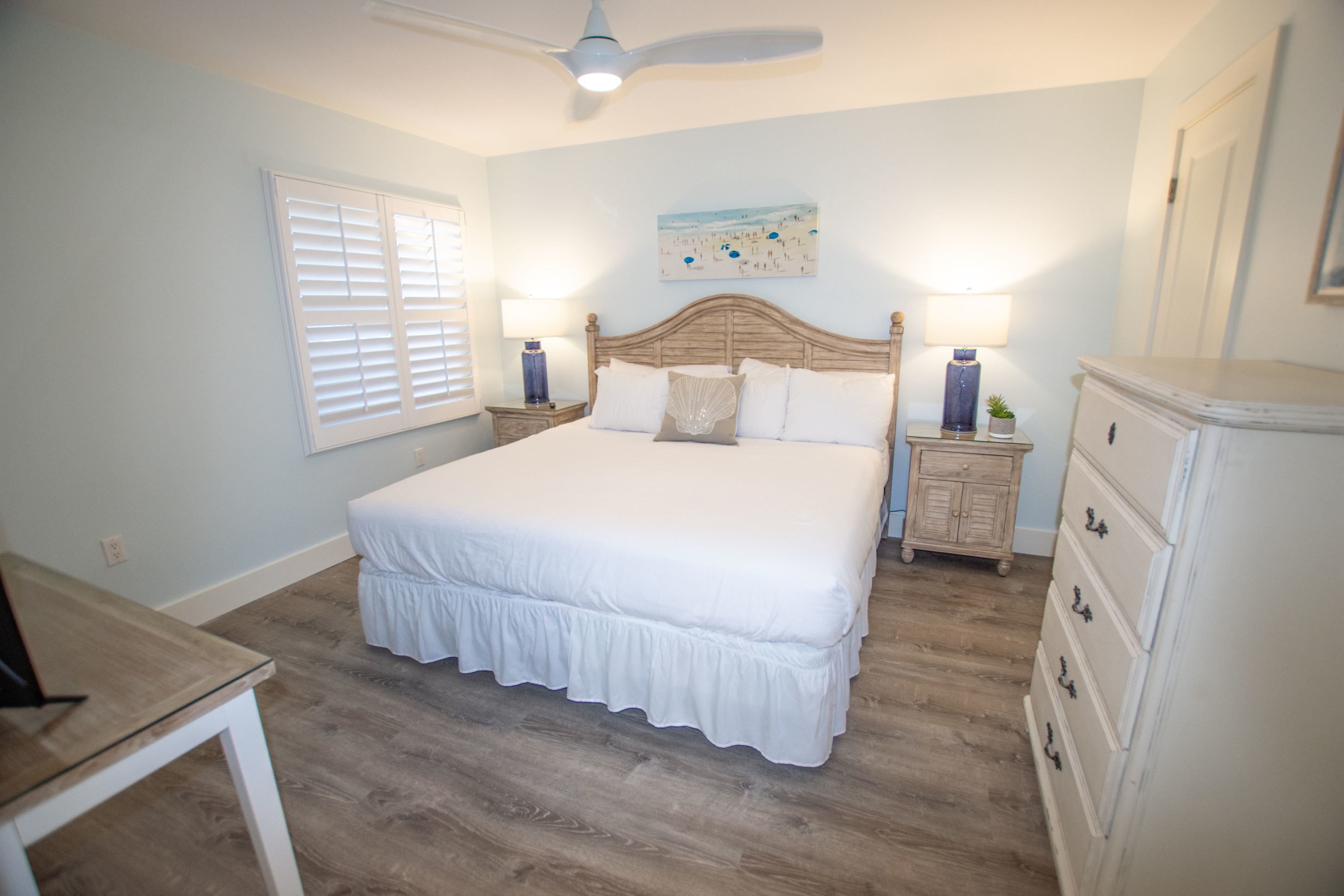 A bedroom with a large bed, two bedside tables with lamps, a ceiling fan, a window with blinds, and a dresser.