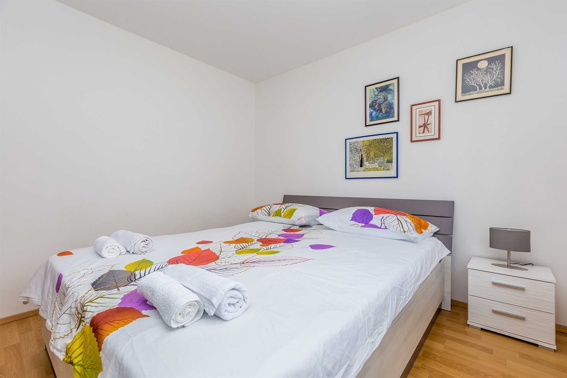 A neatly made bed with a floral-patterned bedspread and rolled towels, a bedside table with a lamp, and three framed artworks on the wall.
