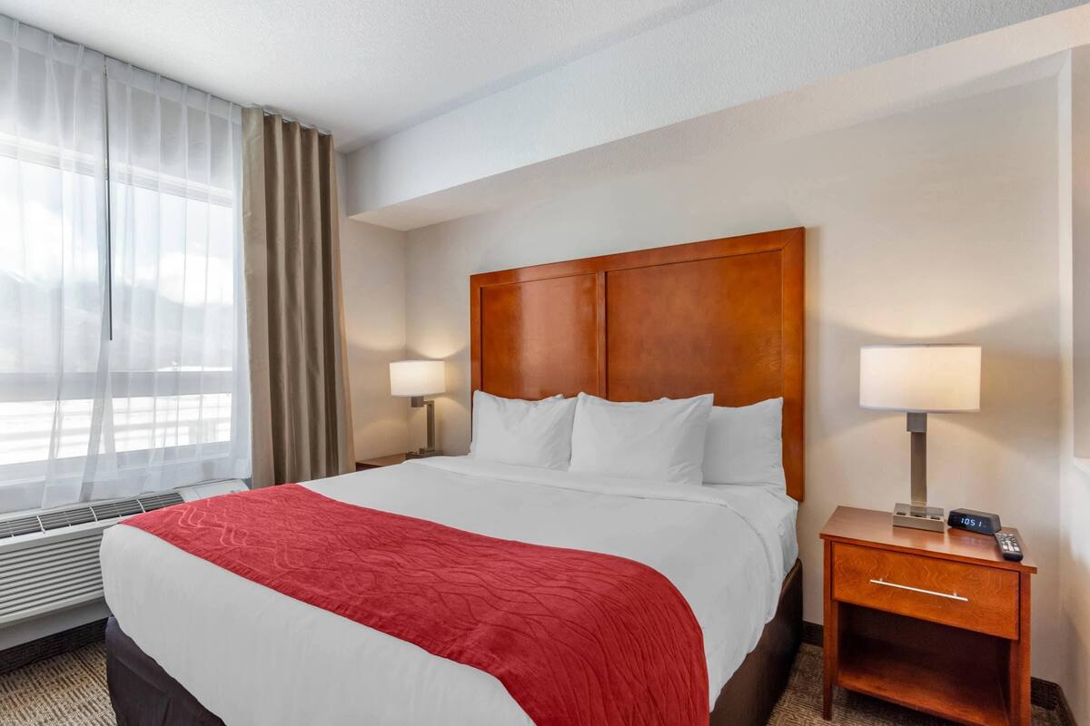 A hotel room with a large bed, a wooden headboard, two bedside tables with lamps, and a window with curtains.