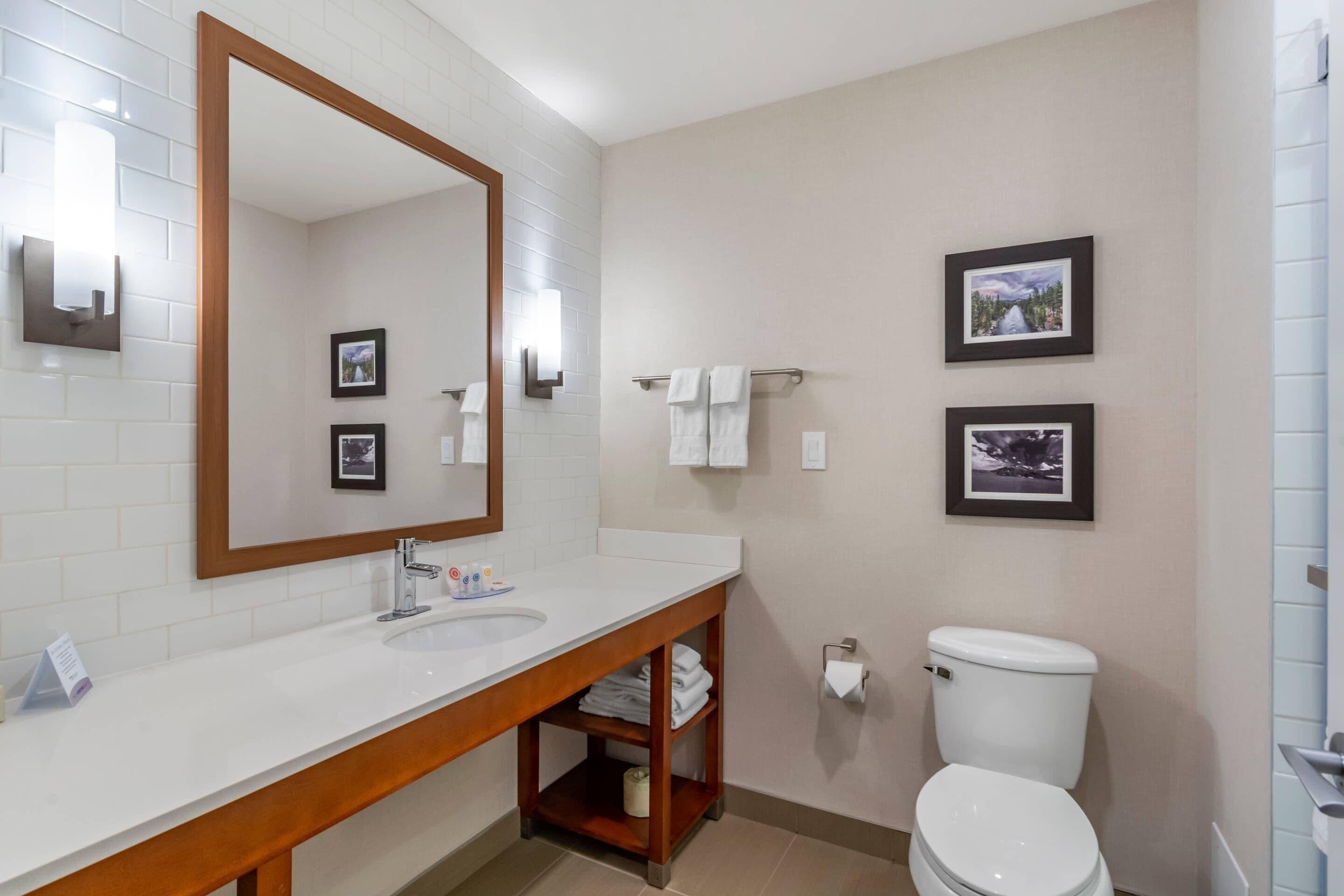A modern bathroom with a white sink, wooden vanity, a large mirror, two framed pictures on the wall, and a white toilet.