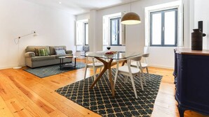 A modern living room with a glass dining table, white chairs, a gray sofa, and a patterned rug.