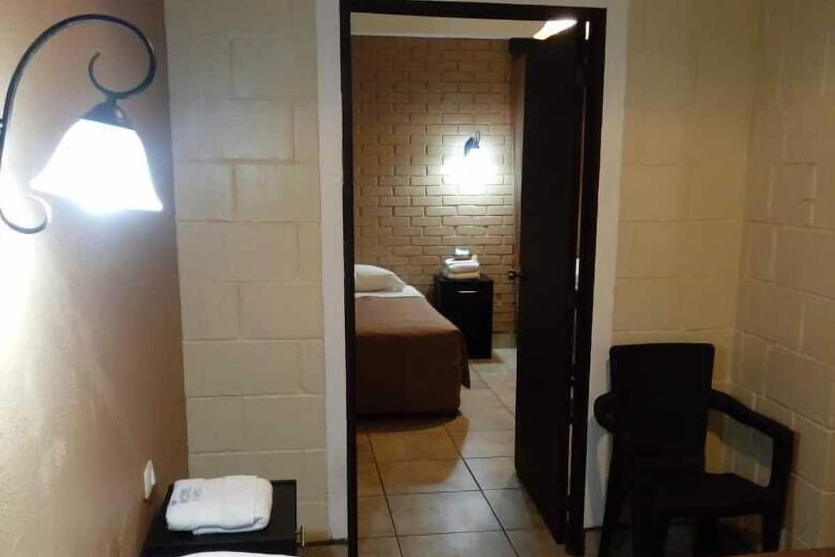 A hotel room with a bed, a chair, a nightstand, and a brick wall.