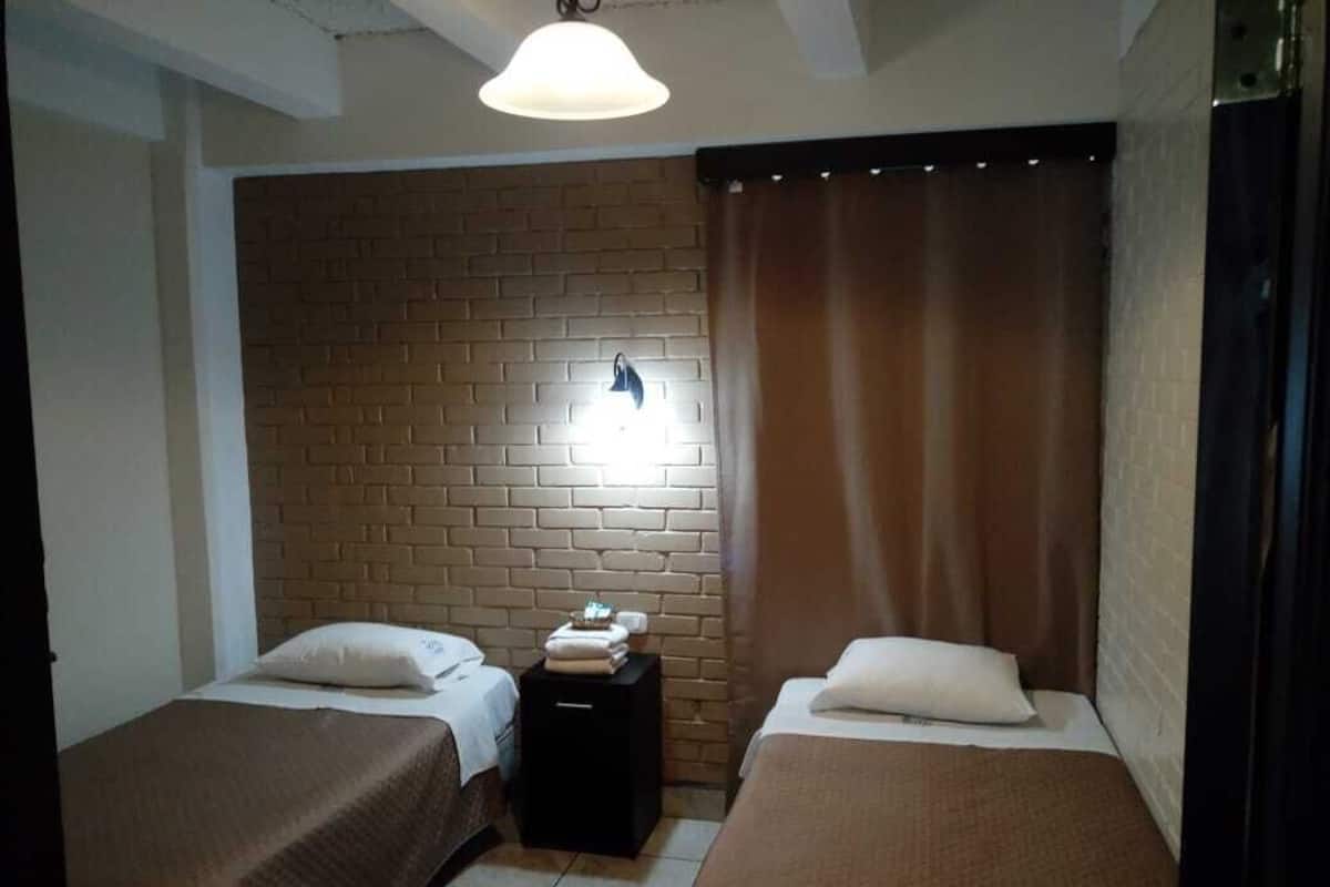 A room with two beds, a brick wall, a small table with a lamp, and a curtain.