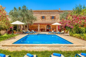 A pool area with lounge chairs and umbrellas, a stone building with a tiled roof, and lush greenery.