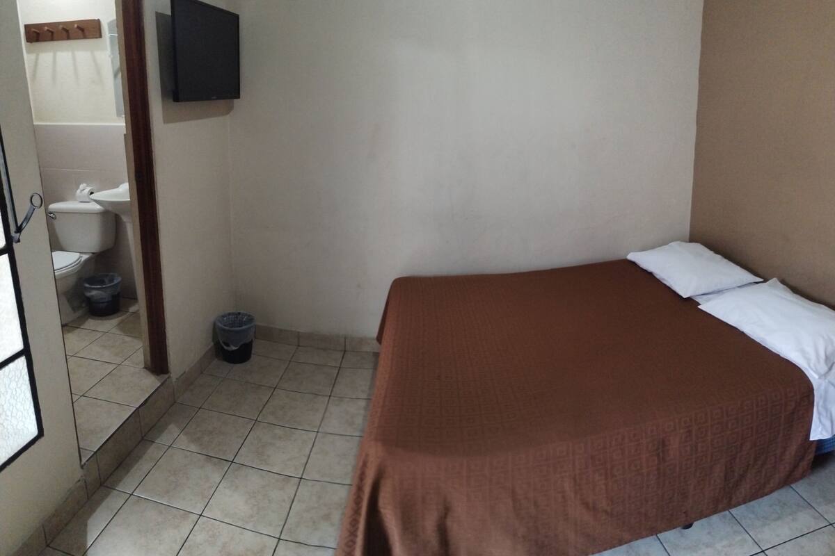 Ein kleines, schlichtes Zimmer mit einem Bett, einem an der Wand befestigten Fernseher, einem Waschbecken und einer Toilette.