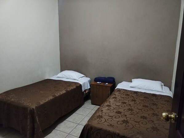 A small, dimly lit room with two single beds, a wooden nightstand, and a suitcase.
