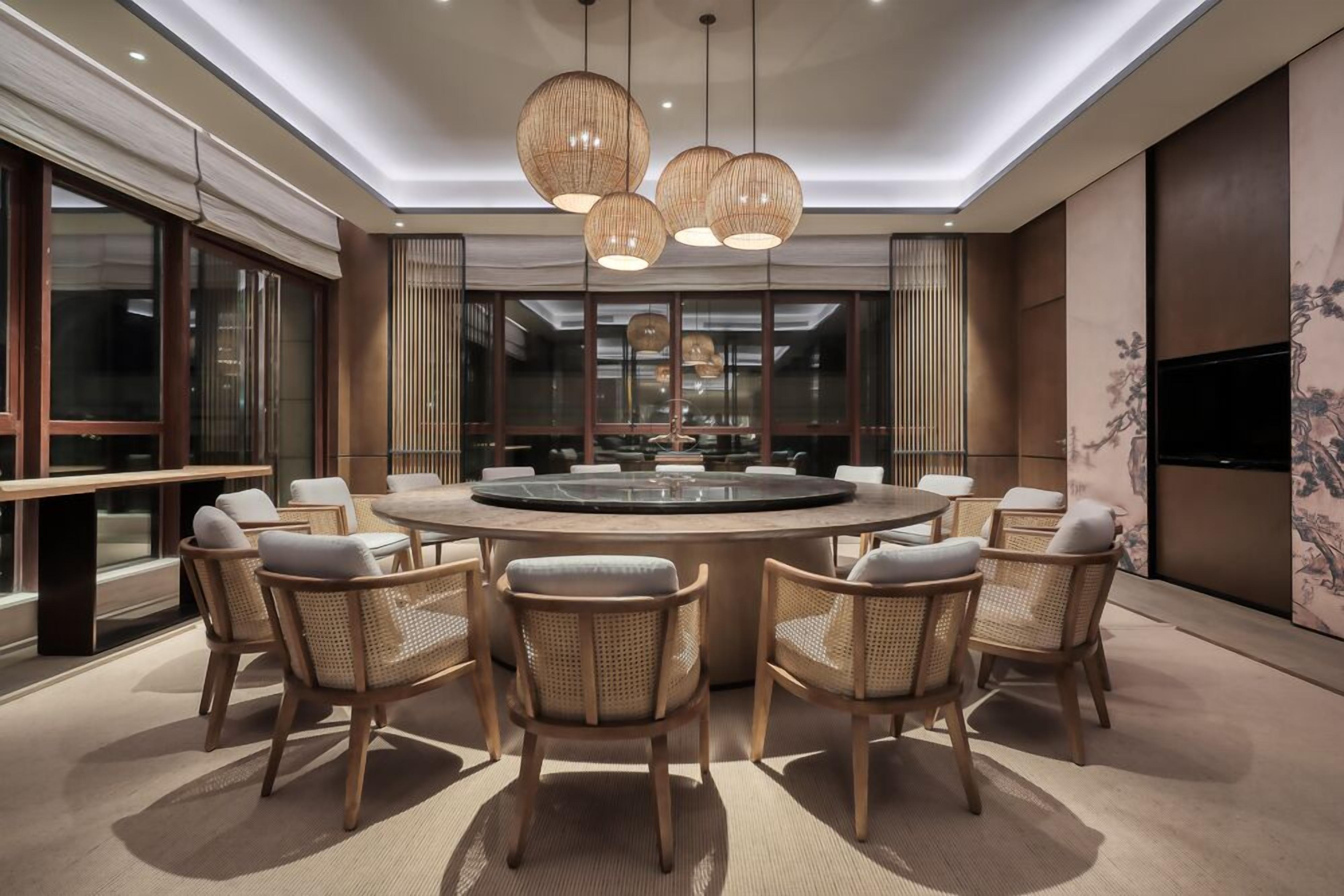 A modern dining area with a round table surrounded by wicker chairs, overhead lighting, and a large window with blinds.