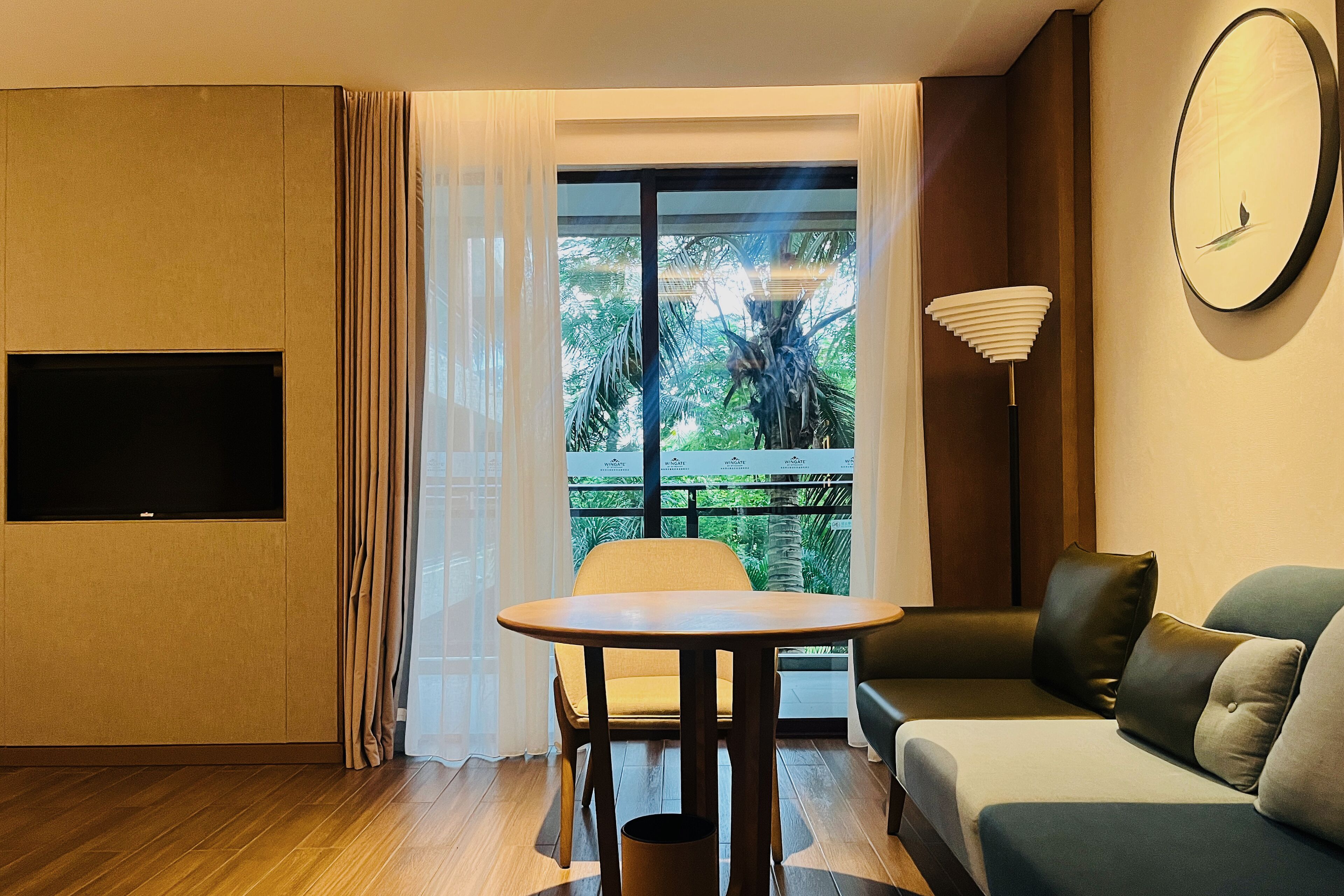 A modern hotel room with a flat-screen TV, a round table, a sofa, and a balcony with a view of greenery.