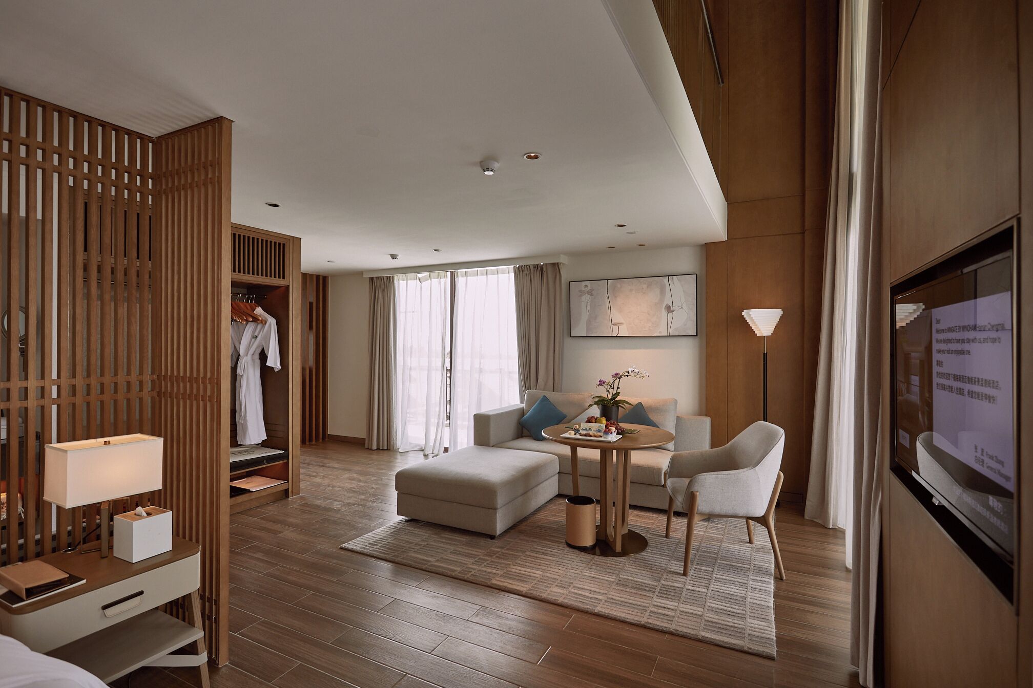 A modern hotel room with a wooden wardrobe, a sofa, a small table with a vase, and a TV mounted on the wall.