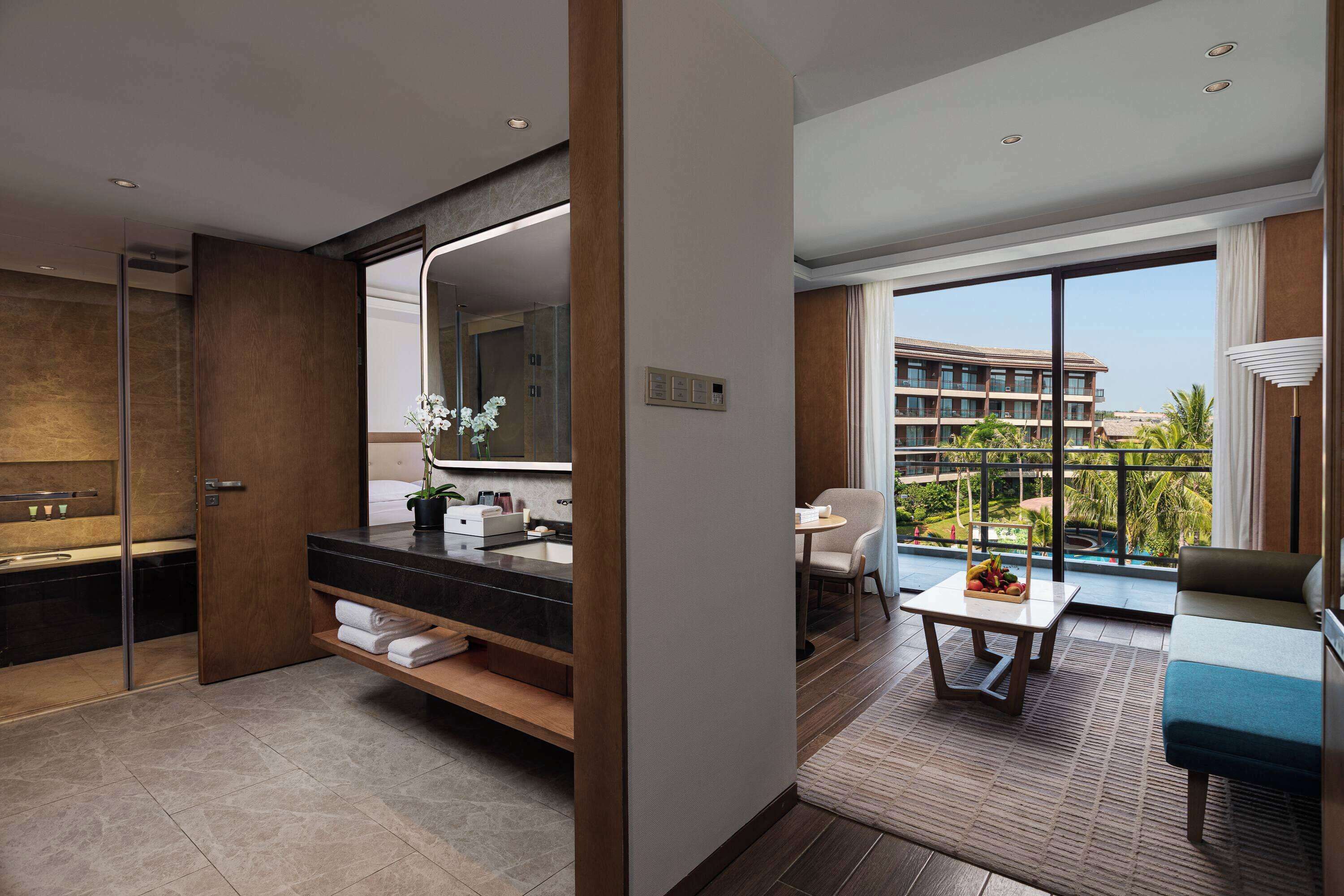 A modern hotel room with a bathroom, a balcony with a view of the hotel and palm trees, a seating area with a table and chairs, and a large mirror.