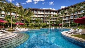 Zona de piscina de hotel con varios niveles de balcones, palmeras y sillas de descanso.