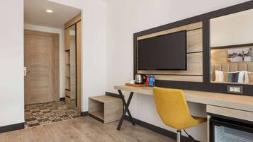 A modern hotel room with a wooden floor, a desk with a chair, a flat-screen TV, and a minibar.