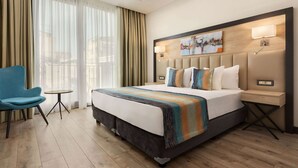 A hotel room with a large bed, a blue armchair, a small table, and a painting on the wall. - Ramada Encore By Wyndham Istanbul Basin Express (Istanbul)