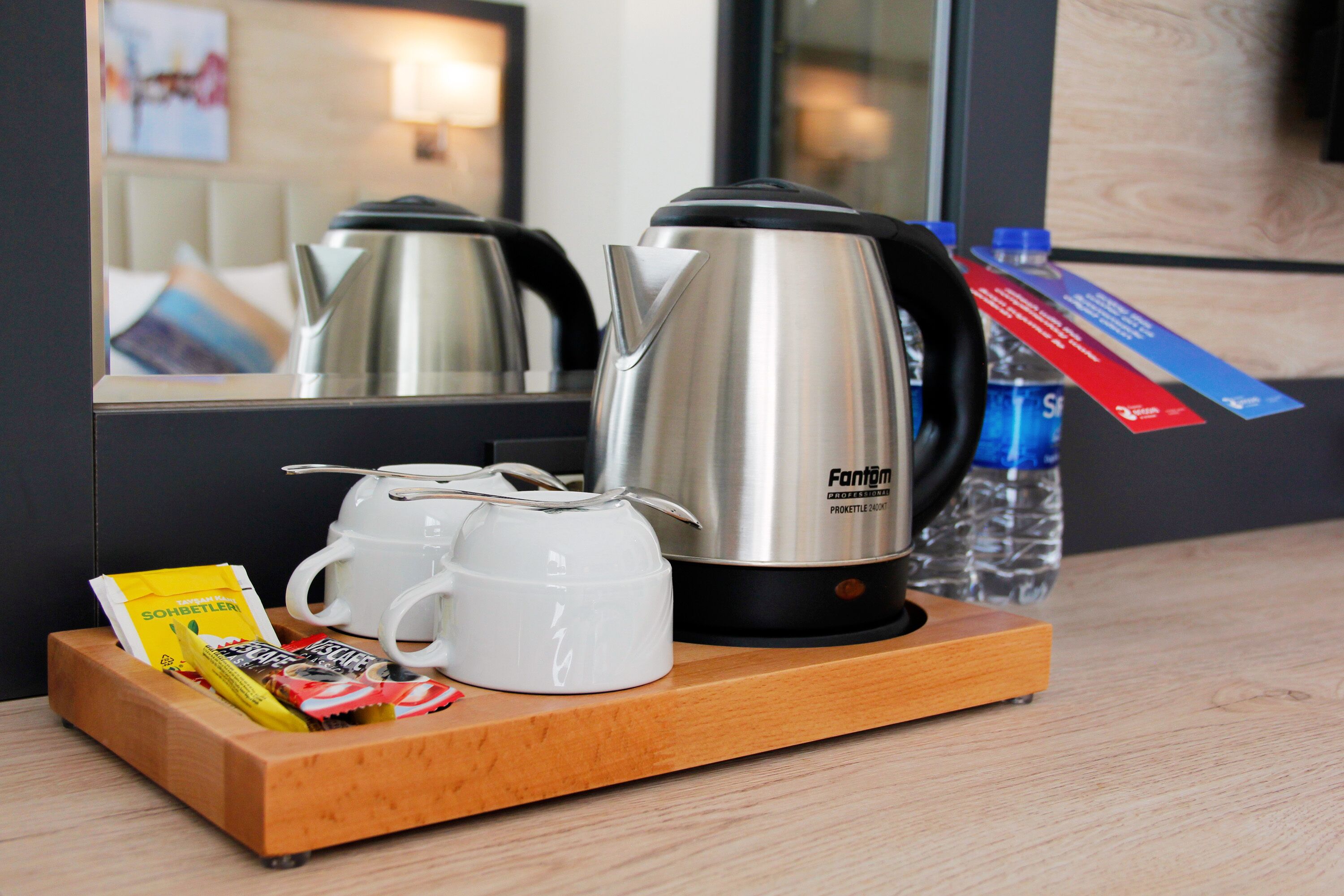 A breakfast tray with a stainless steel kettle, two white mugs, and assorted snacks on a wooden tray.