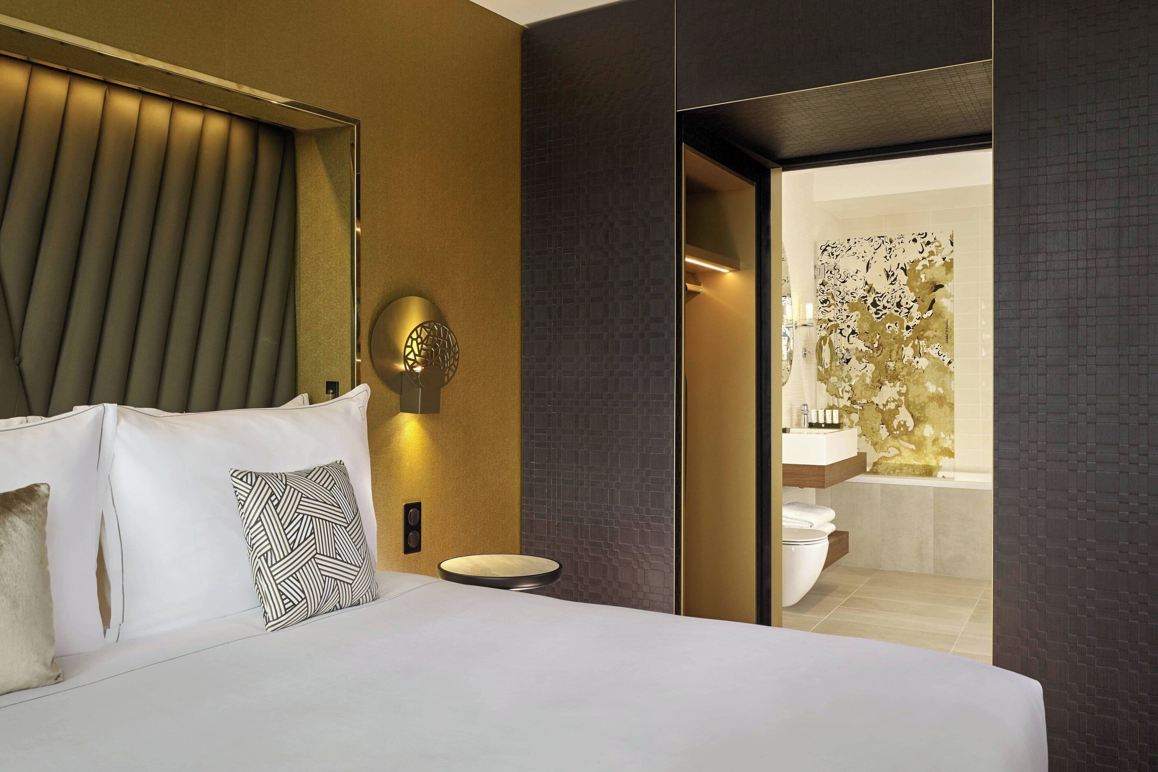 Una habitación de hotel moderna con una cama grande, una mesita de noche, una lámpara de pared y vistas al baño.