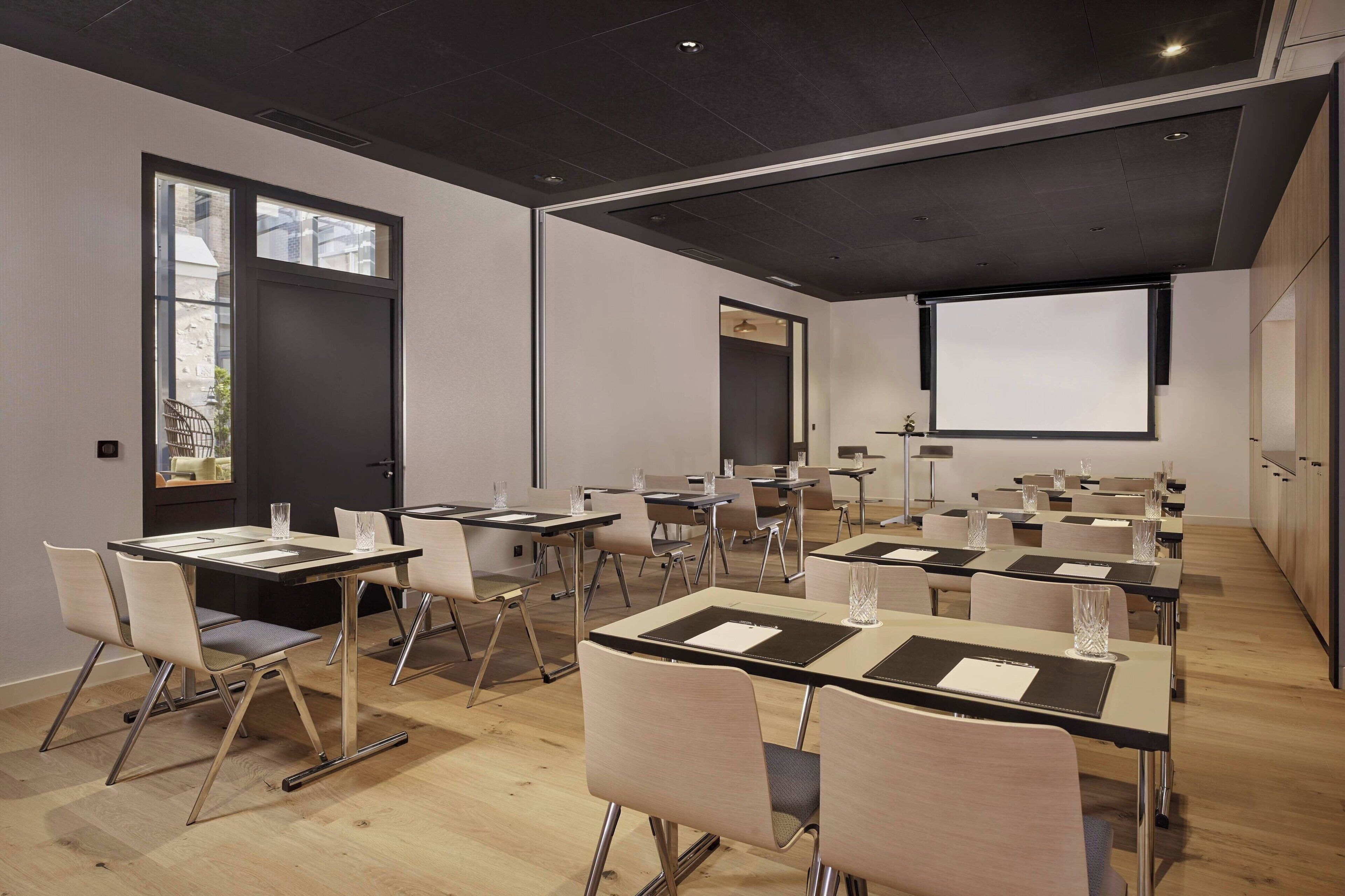 Un moderno salón de conferencias con varias mesas y sillas, una pantalla de proyector y un ventanal grande.