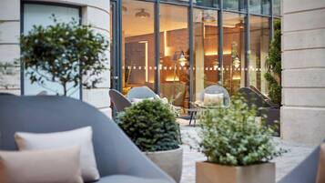 Een terras met moderne meubels, potplanten en een zicht op het interieur door grote ramen.