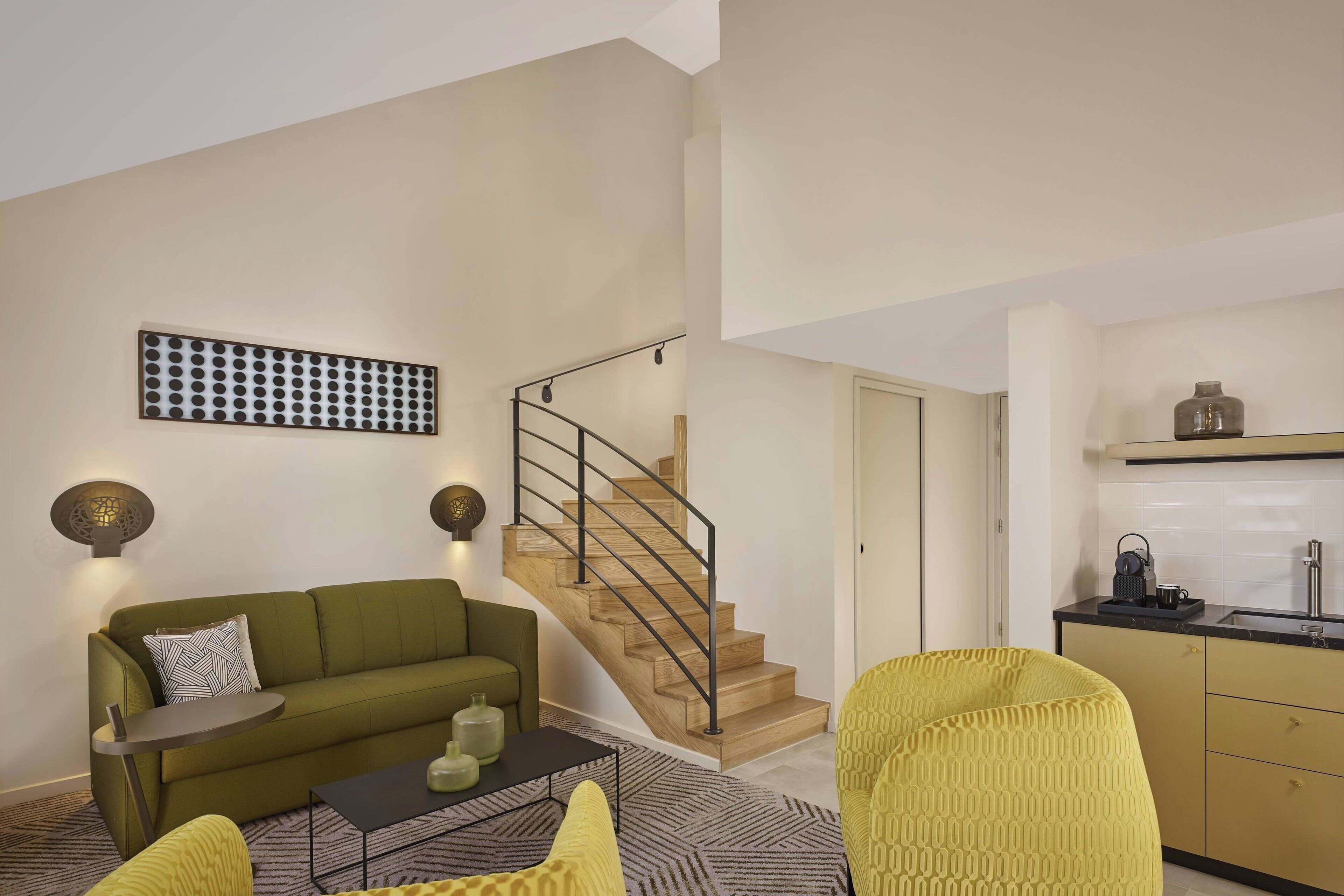 Un salon moderne avec un canapé vert, une petite table ronde et un escalier menant à un espace cuisine.