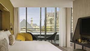 A hotel room with a large bed, a TV, a small table, and two chairs. There is a view of a building with a tower through the window. - La Caserne Chanzy Hotel & Spa, Autograph Collection (Reims)