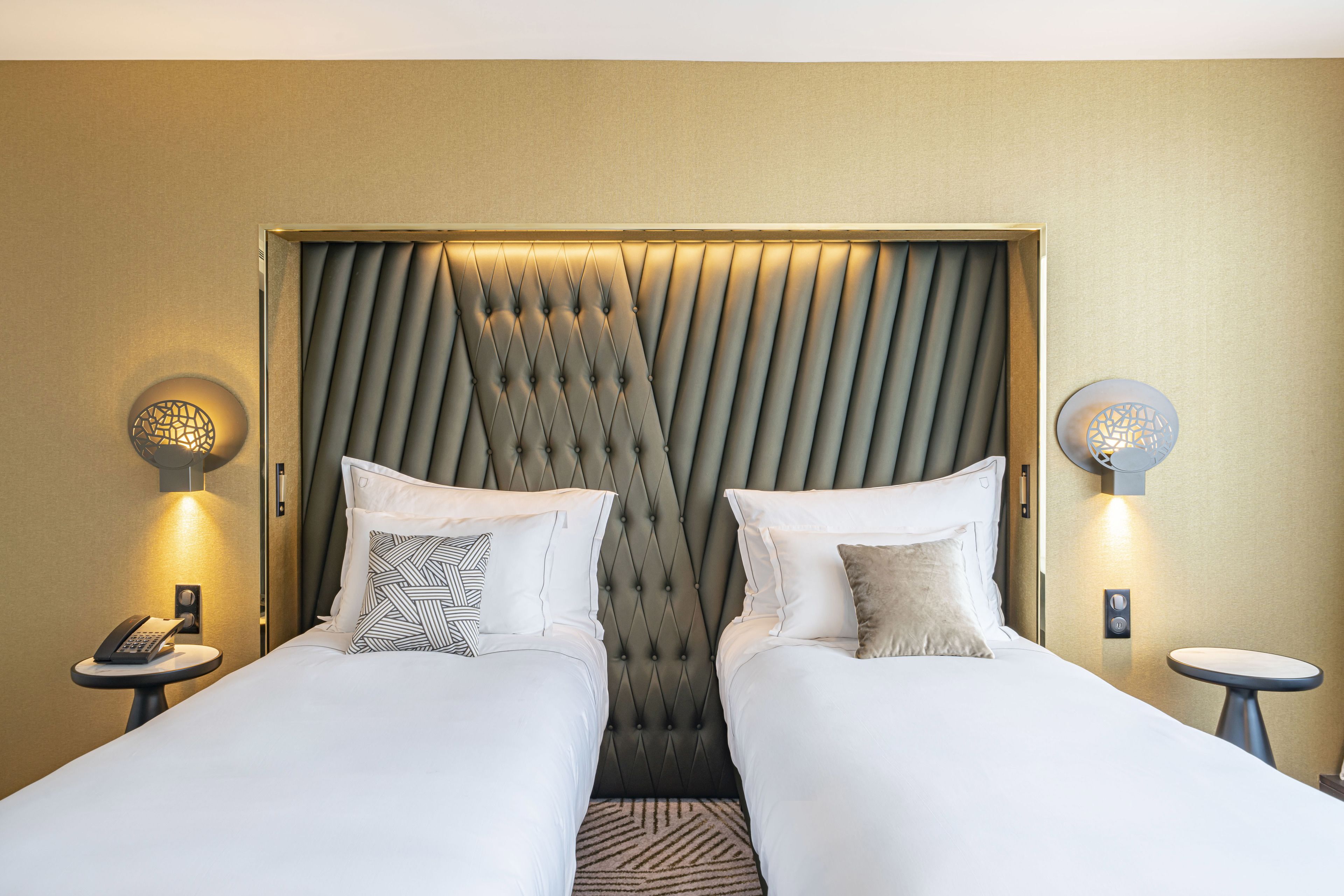 Habitació d'hotel amb dos llits, un reposacapses amb textura, petites làmpades, una taula petita i un telèfon.