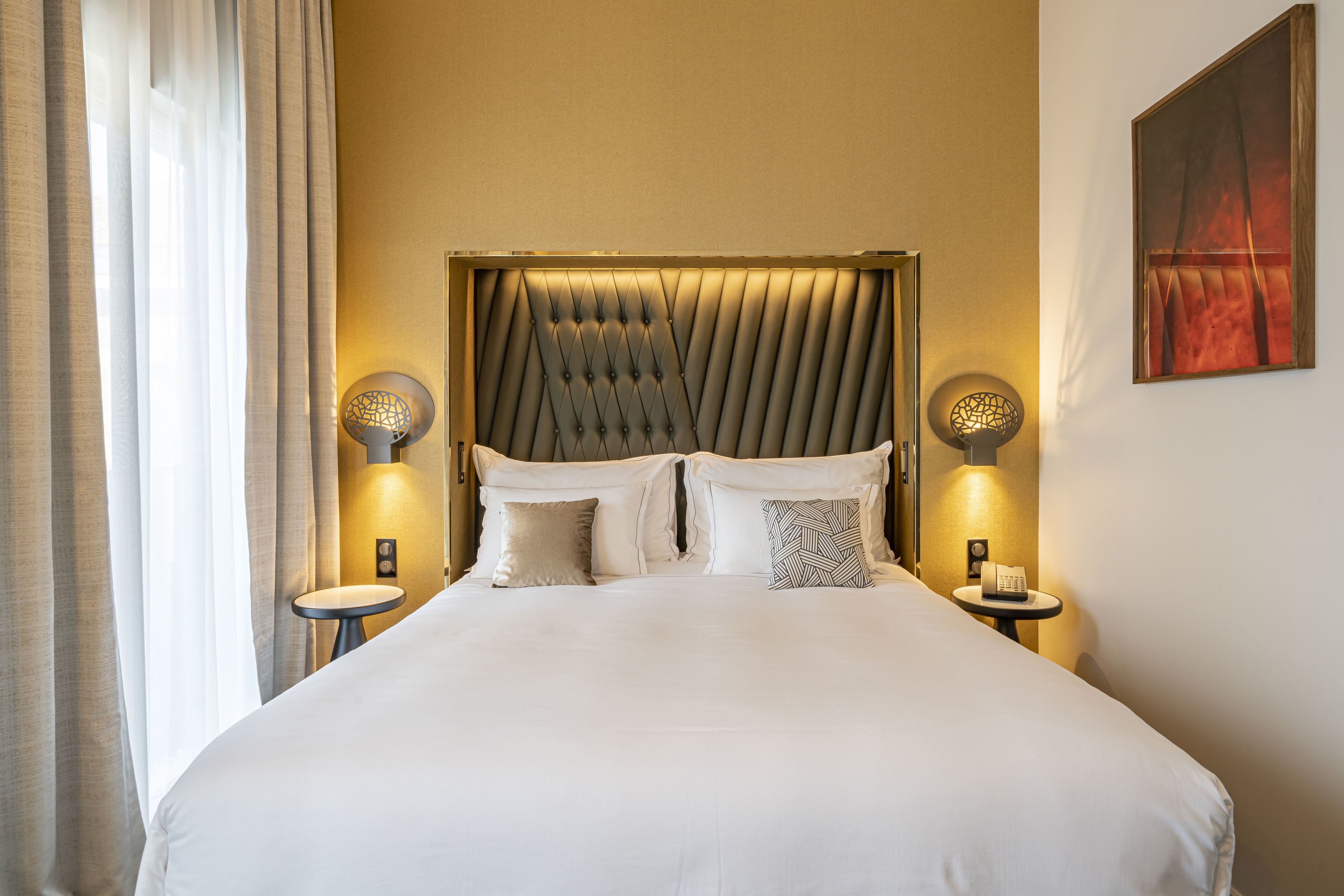 A large bed with a tufted headboard, two bedside tables with lamps, and a painting on the wall.