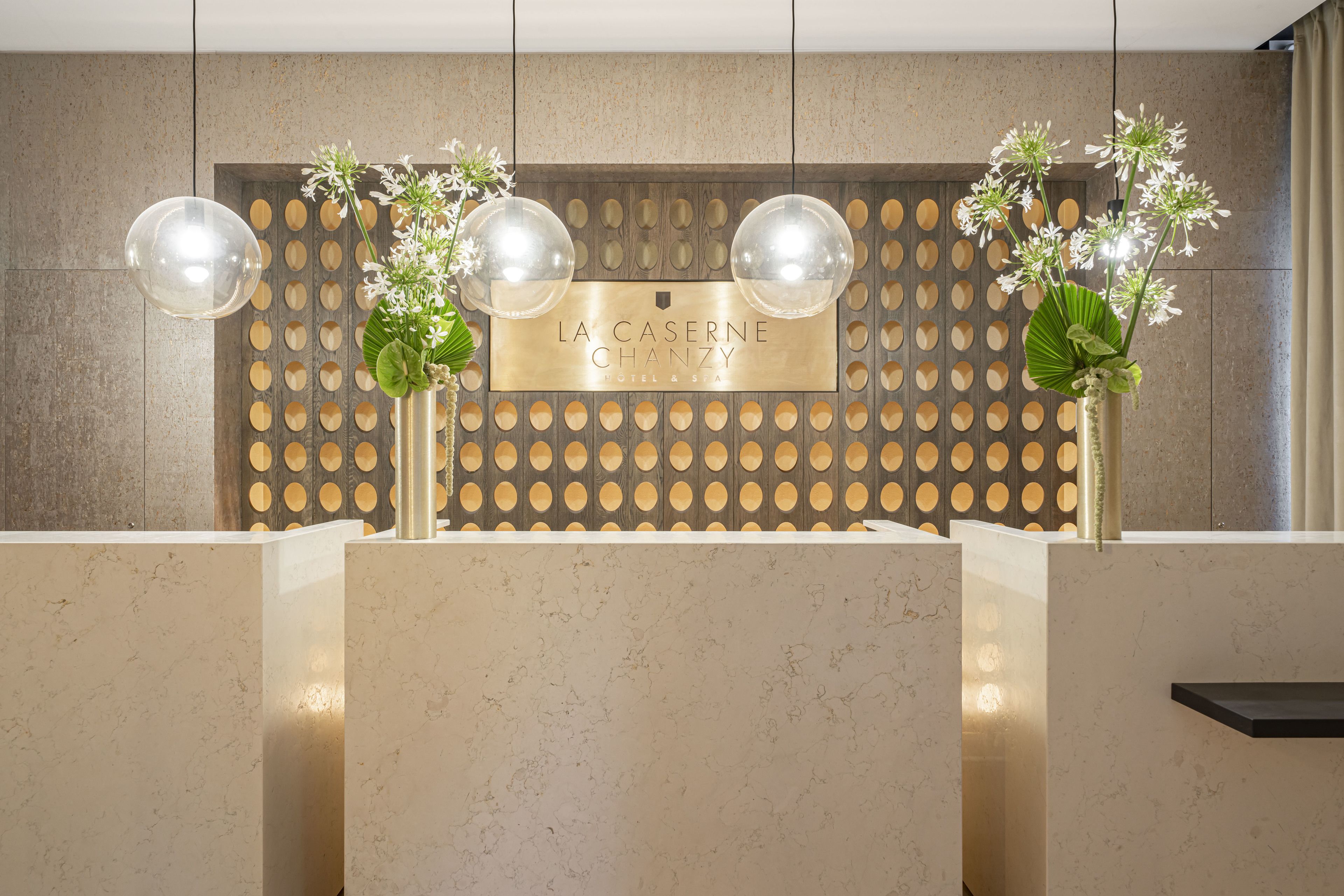 Un espace d’accueil doté d’un comptoir en marbre, d’une enseigne murale indiquant « HÔTEL ET SPA LA CASERNE CHANZY », et de deux vases ornés de fleurs.