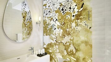 Ein Badezimmer mit rundem Spiegel, weißem Waschbecken und einer Wand mit goldenem und weißem abstrakten Dekor.