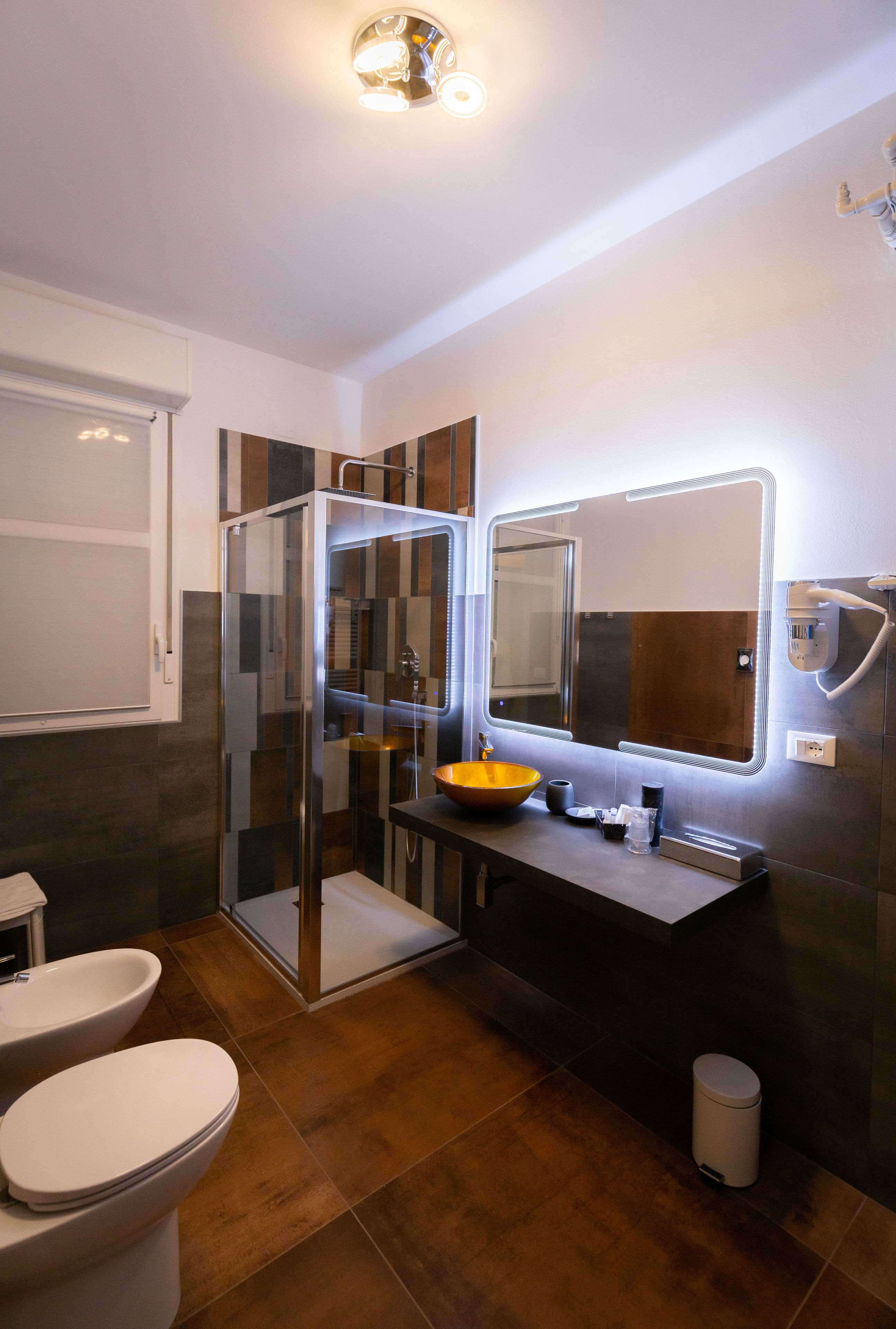 Un bagno moderno con cabina doccia in vetro, uno specchio grande dotato di illuminazione integrata, un mobile bagno in legno e un water bianco.