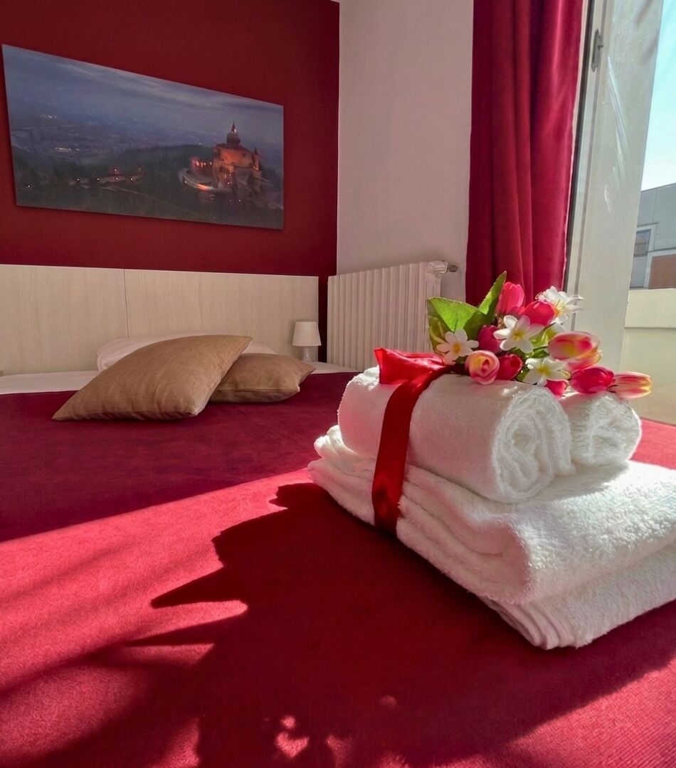 Une chambre d’hôtel avec un tapis rouge, un tableau au mur, un lit avec une serviette rouge et blanche, et une fenêtre avec des rideaux.