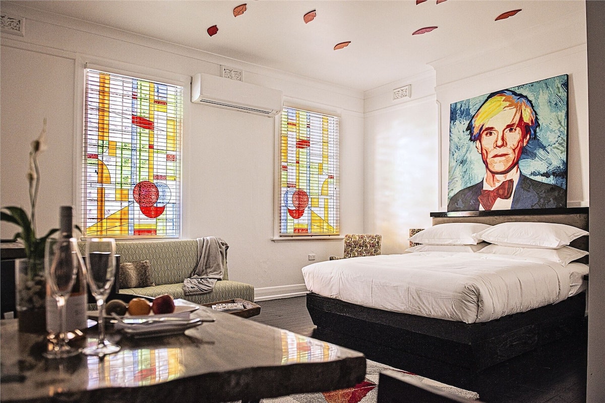 A hotel room with a bed, a chair, a table with glasses and fruits, and two windows with stained glass.