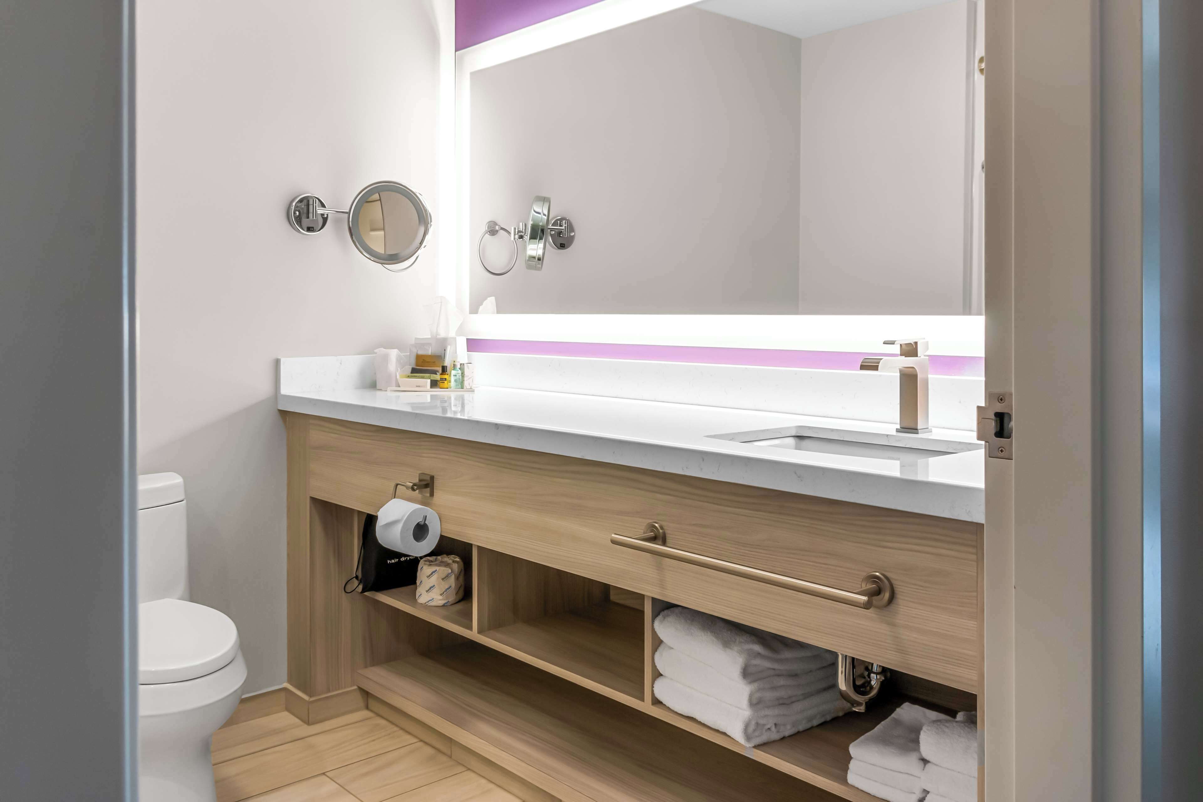 A modern bathroom with a white sink, mirror, and toilet. There are towels and toiletries on the counter and shelves.