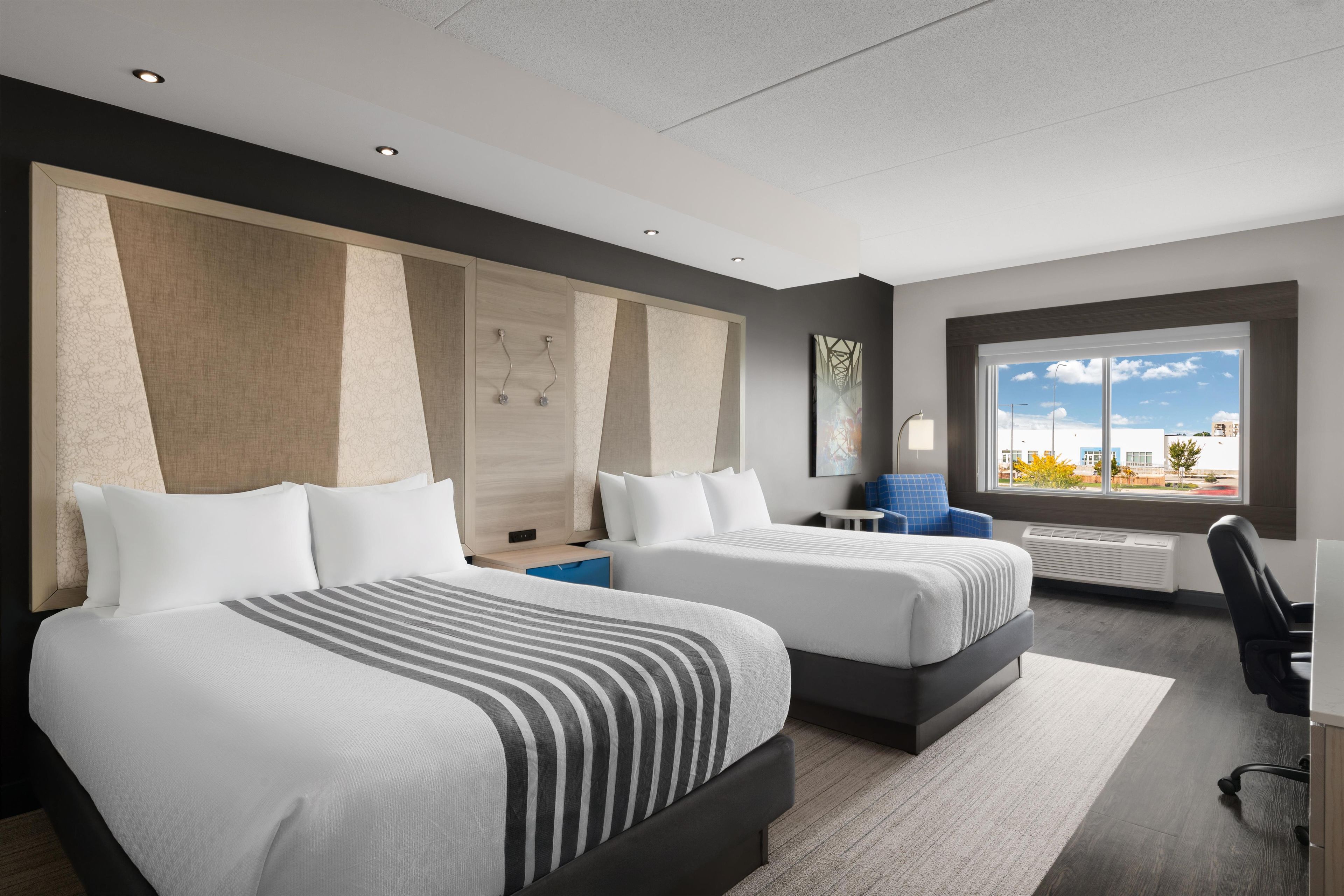 Moderni hotellihuone, jossa on kaksi sänkyä, työpöytä ja suuri ikkuna, josta avautuu näkymä kaupunkiin.