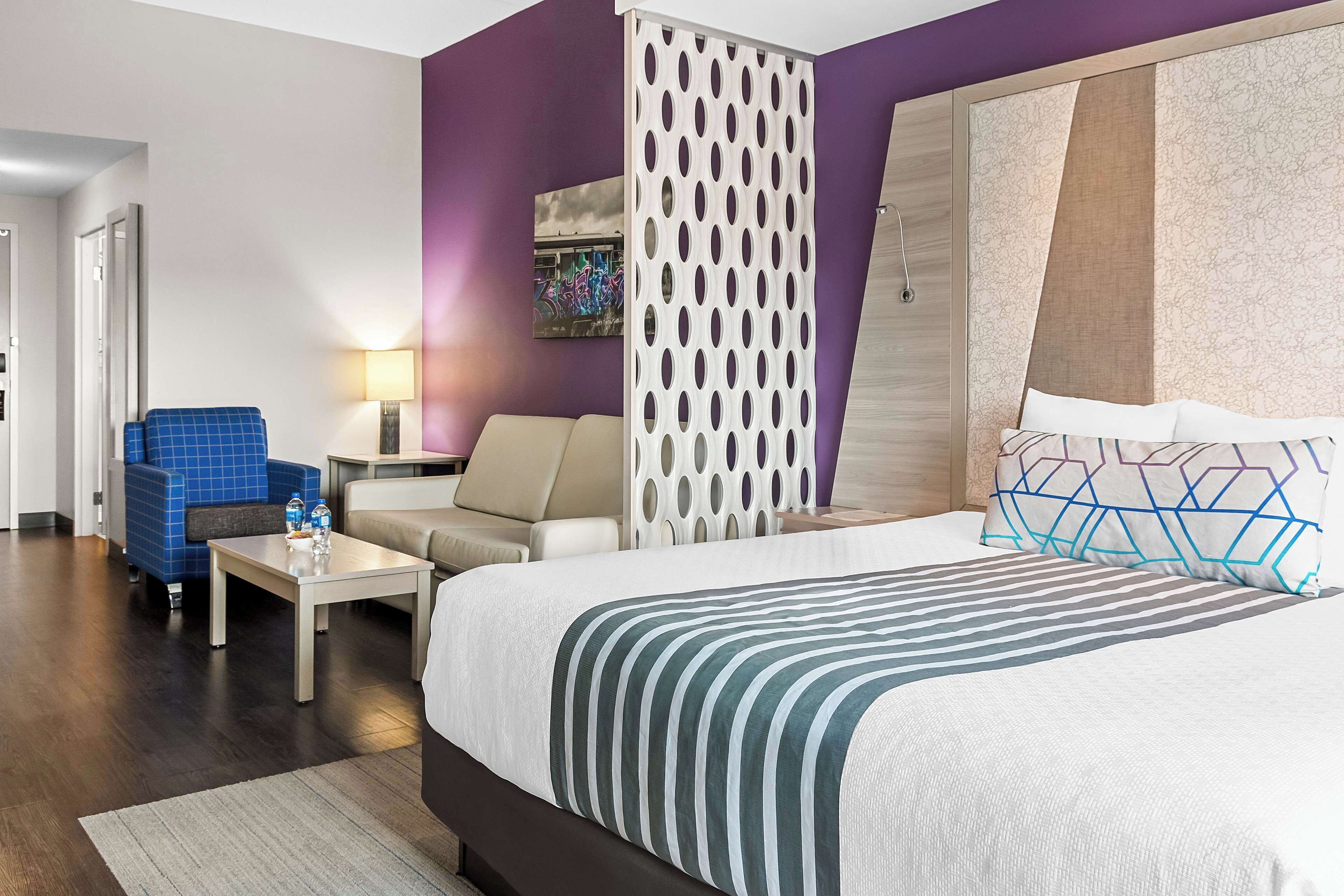 Habitación de hotel con cama, un sillón azul, un sofá beige, un separador blanco y un cuadro en la pared.