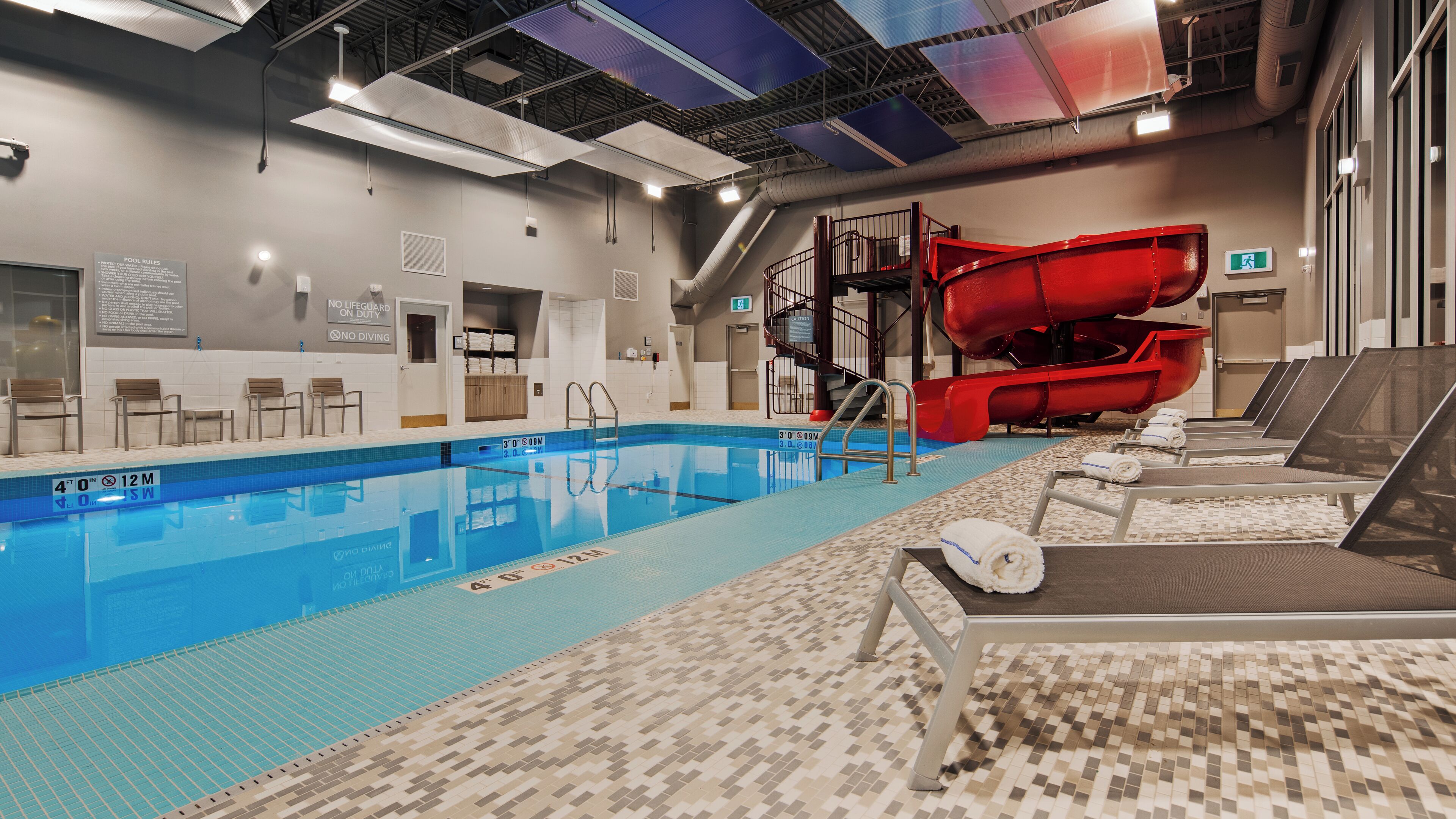 An indoor swimming pool with a red slide, lounge chairs, and a tiled floor.