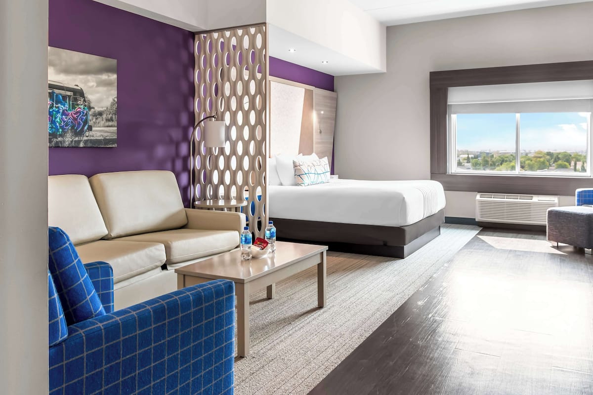 Ein modernes Hotelzimmer mit Bett, Sofa, Couchtisch und Blick auf die Stadt durch ein großes Fenster.