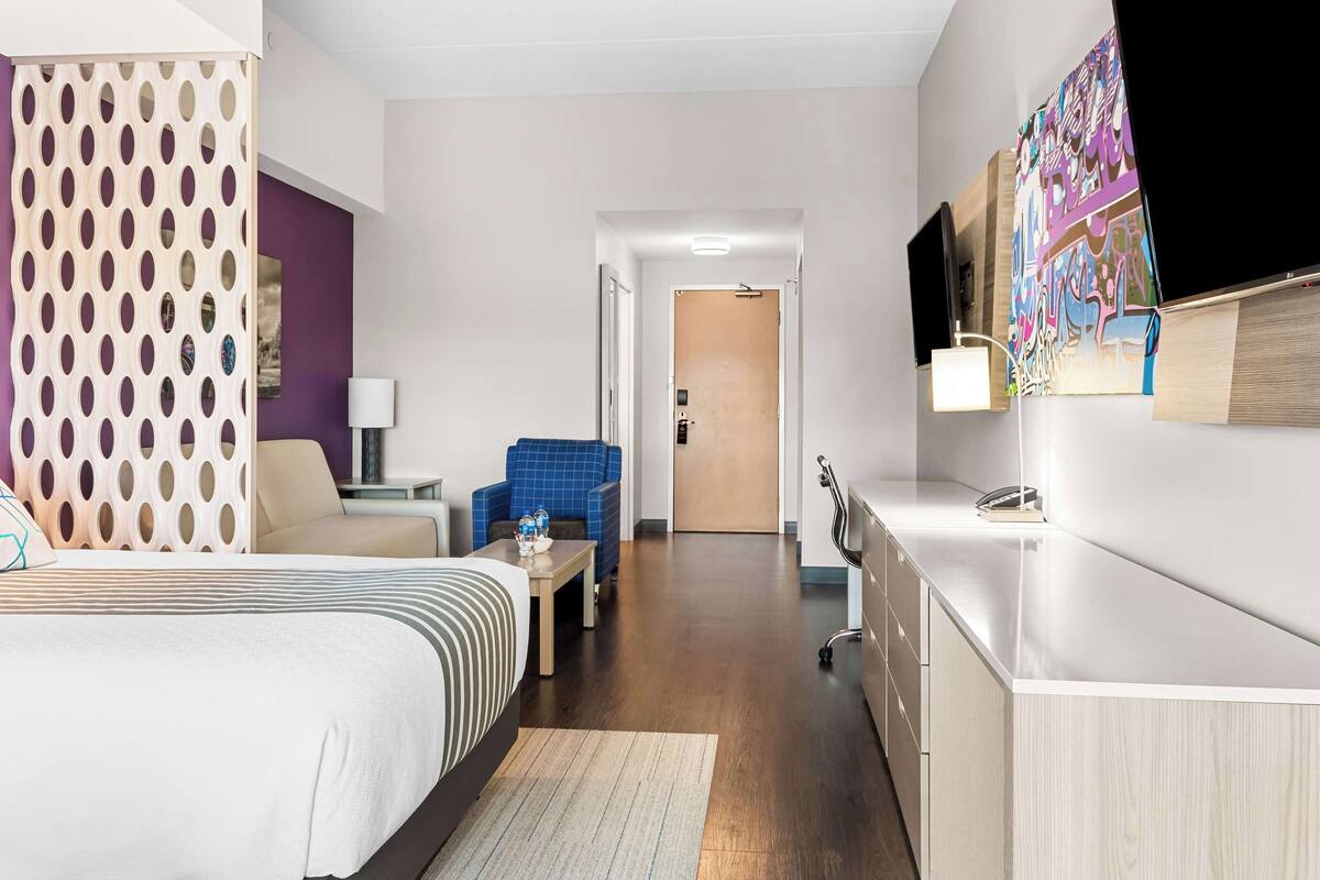 Ein Hotelzimmer mit Bett, Schreibtisch, Stuhl, Fernseher und einer Tür zu einem weiteren Raum.
