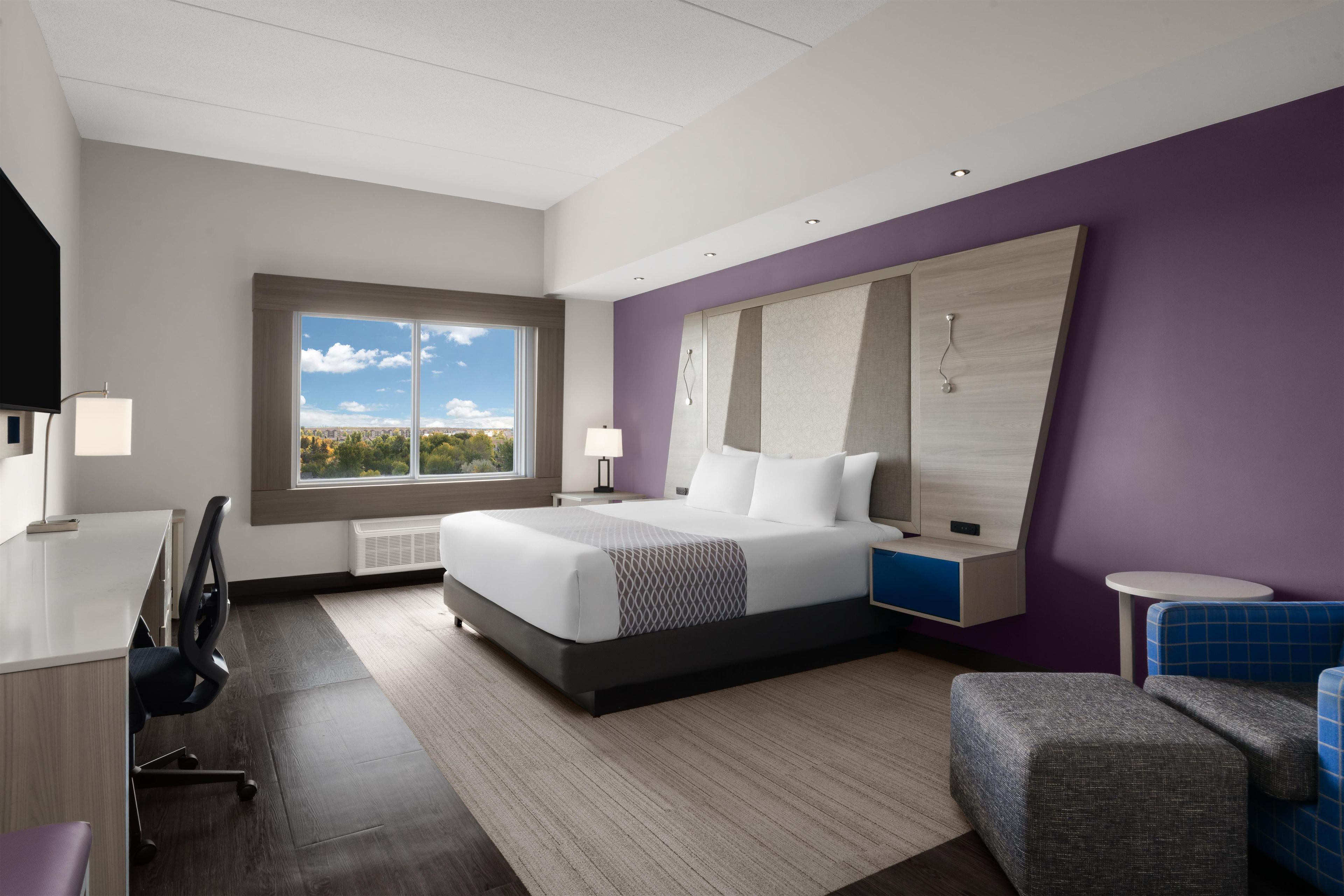 A modern hotel room with a large bed, a desk, a chair, and a view of the cityscape through the window.