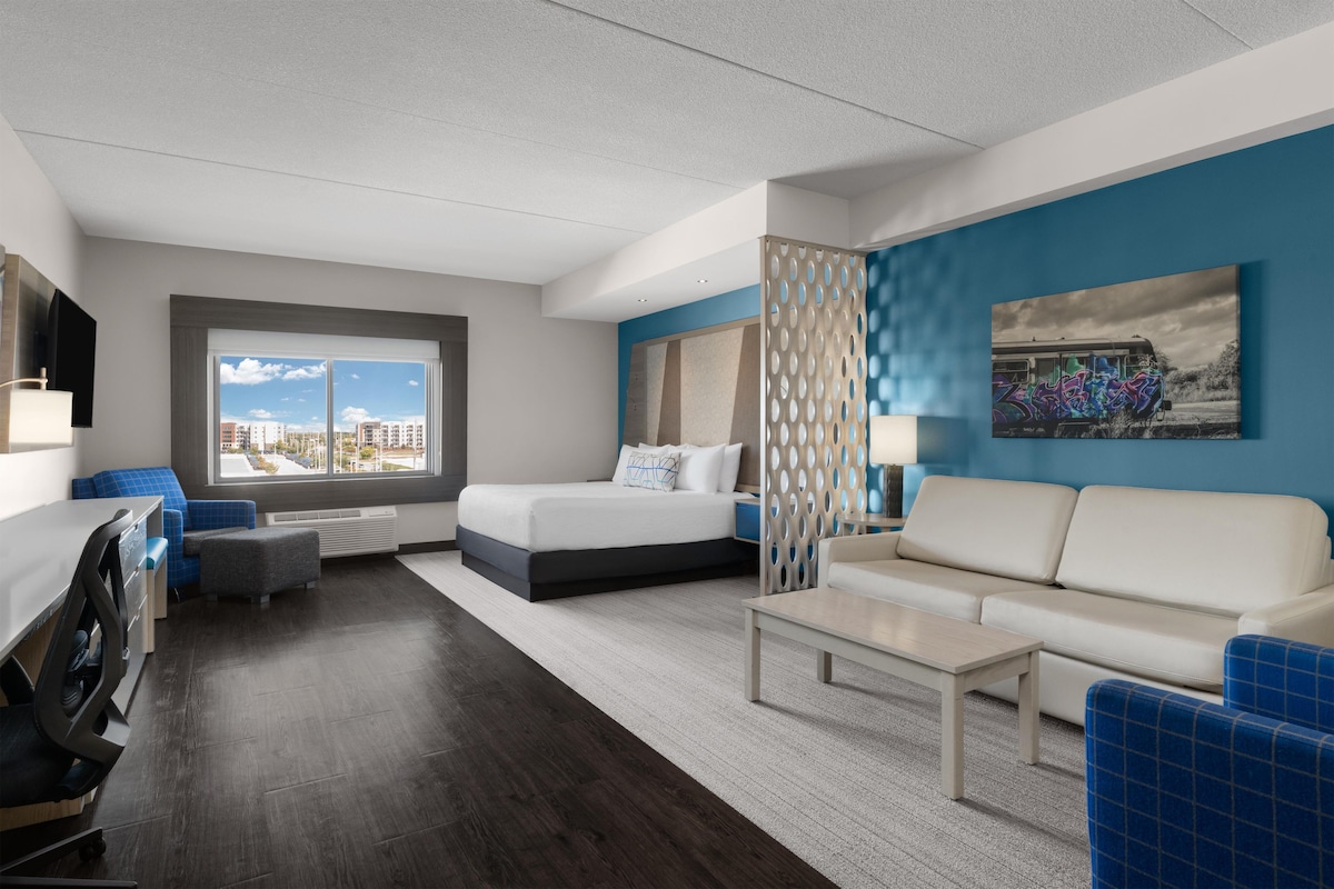 Une chambre d’hôtel moderne équipée d’un lit, d’un canapé, d’un bureau et d’une grande fenêtre donnant sur la ville.