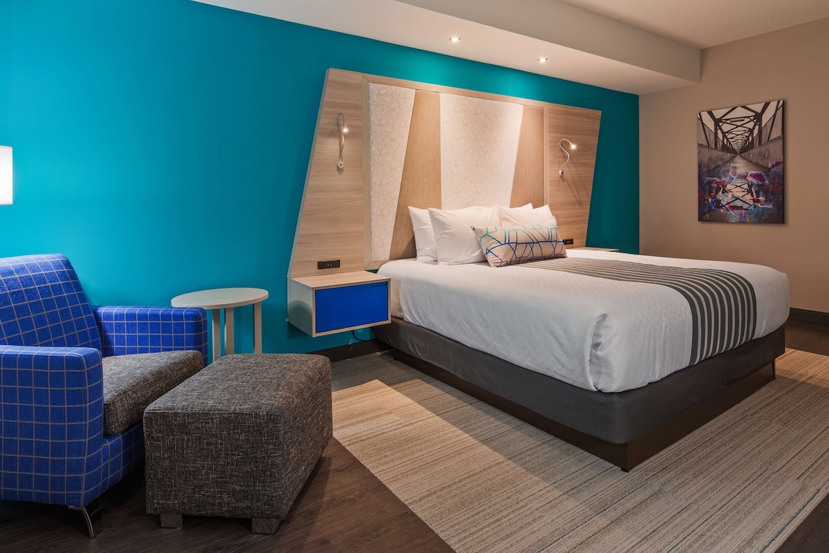 Ein modernes Hotelzimmer mit einem großen Bett, einem blauen Sessel, einem kleinen runden Tisch und einem Nachttisch mit einer Lampe.