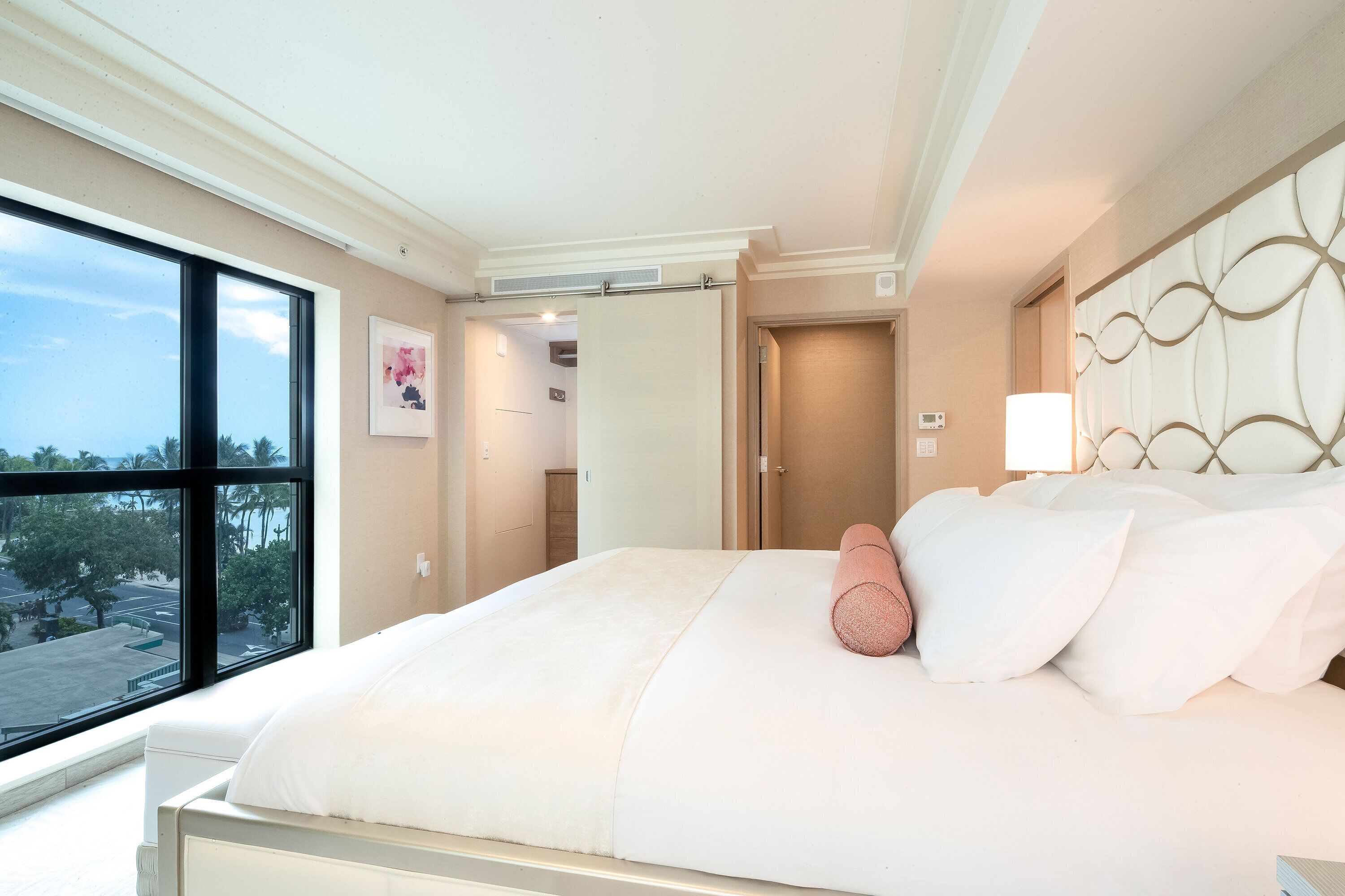 Habitación de hotel con un ventanal grande, cama con ropa de cama blanca, almohada rosa y cabecero decorativo.