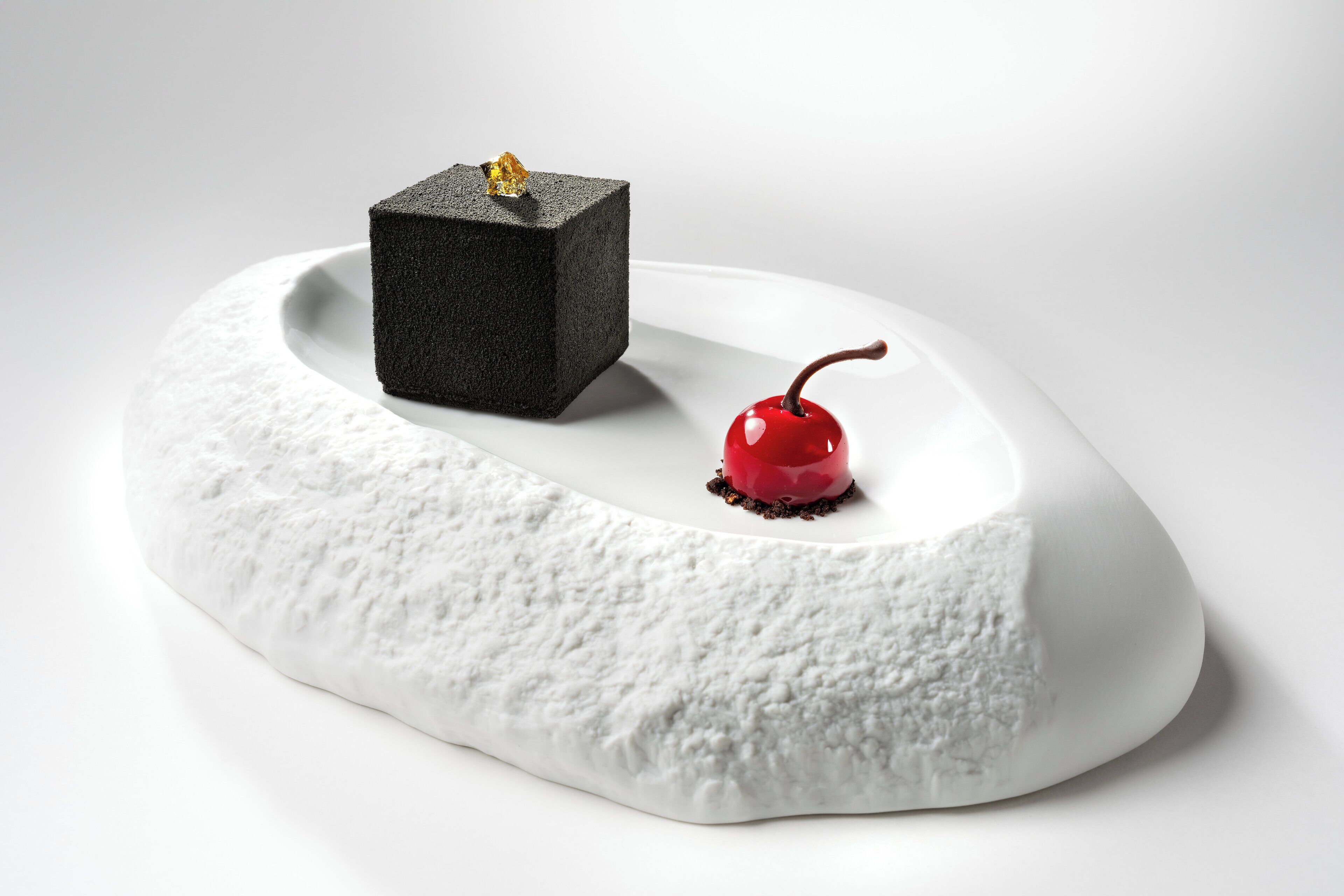 A white, textured surface with a black cube and a red cherry.