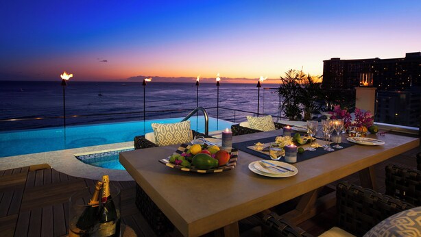 Un área de comedor junto a la piscina con vistas al océano al atardecer, una mesa dispuesta con platos, vasos y un frutero.
