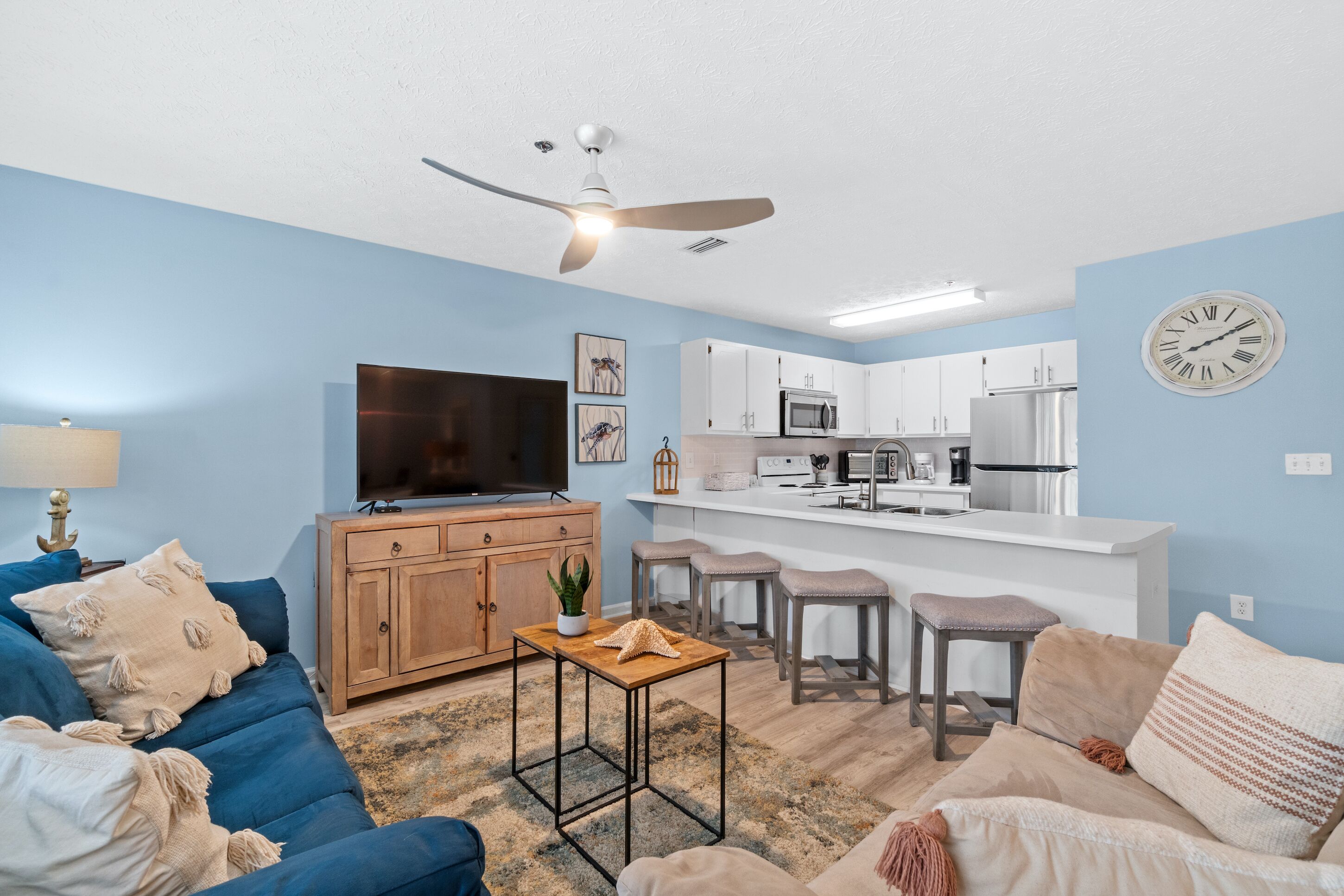 A living room with a blue sofa, a wooden cabinet, a TV, and a kitchen area with white cabinets and a microwave.
