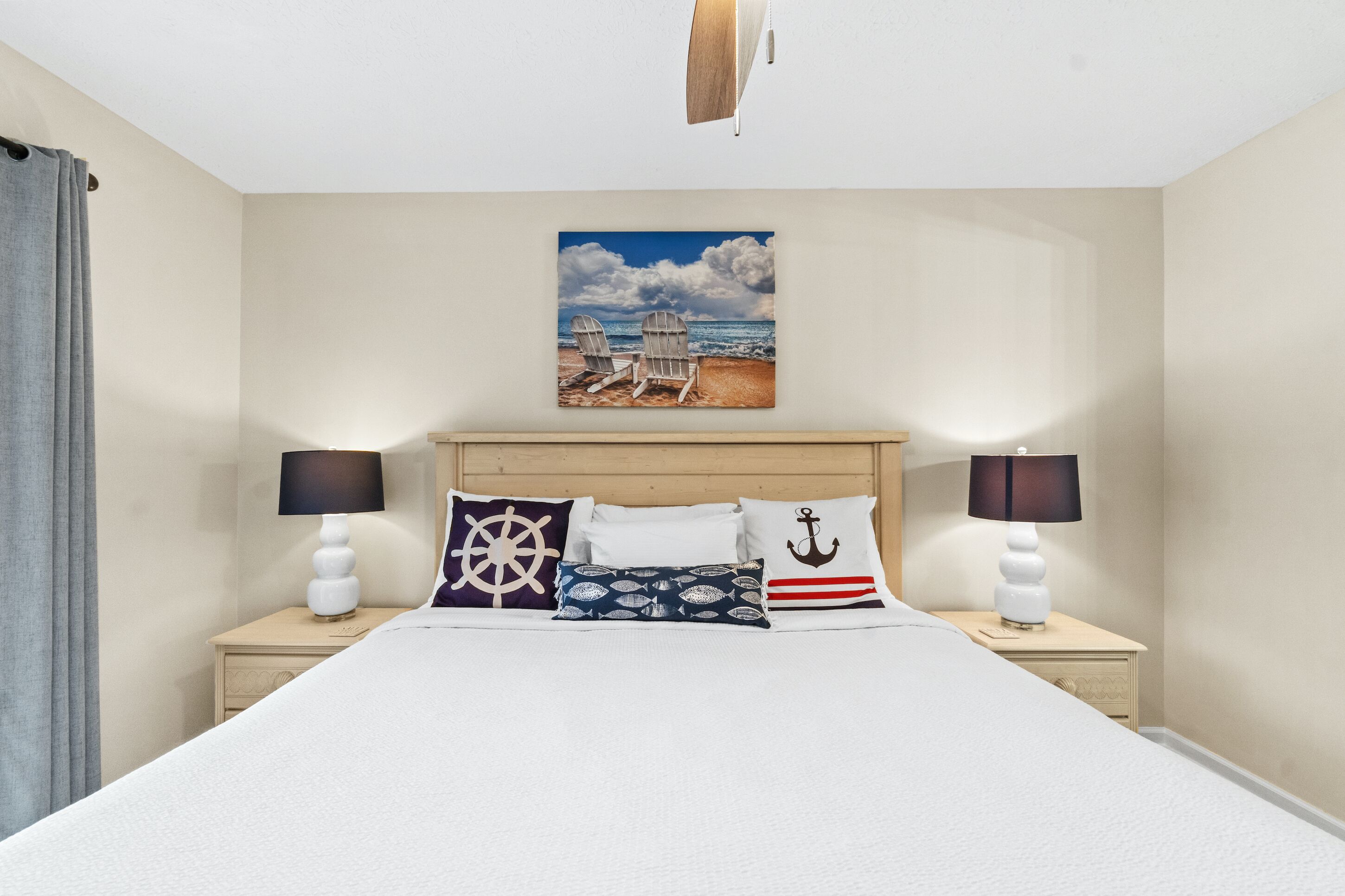 Una cama bien hecha con cabecera, dos mesitas de noche con lámparas y un cuadro con una escena de playa sobre la cama.