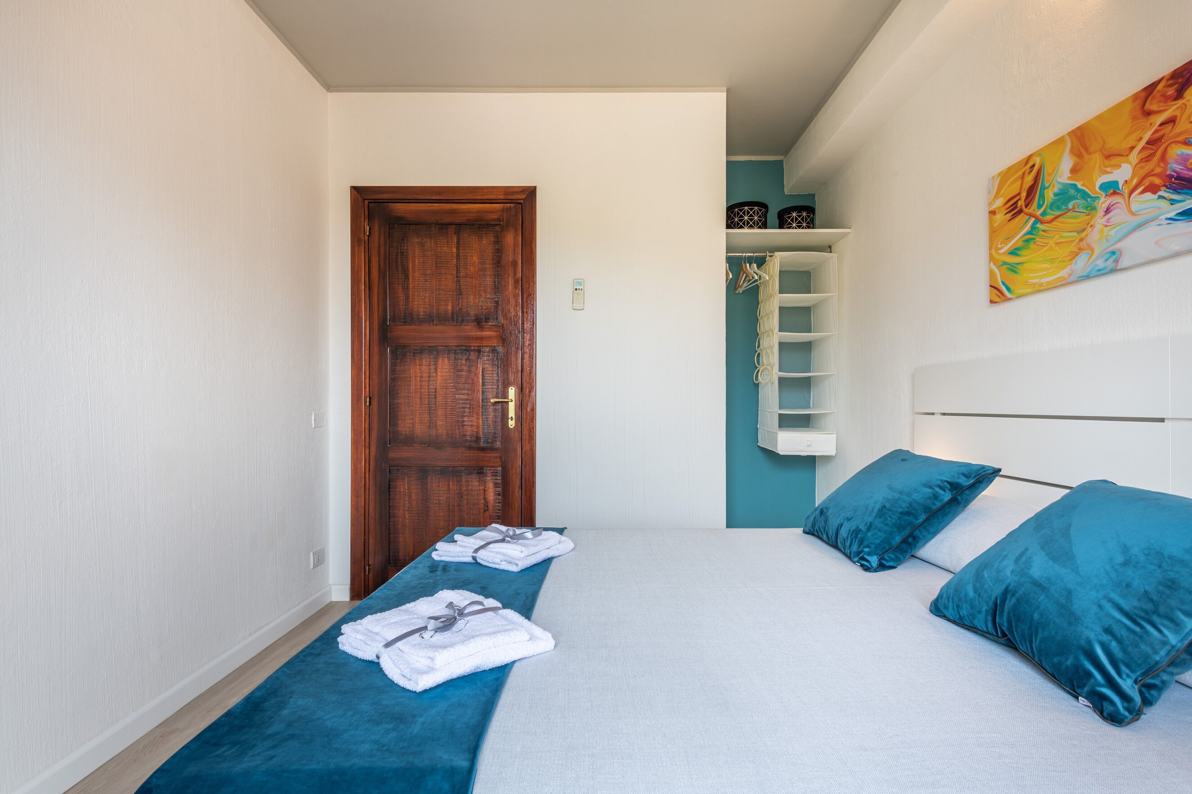A bedroom with a bed, a wooden door, a white wall, and a colorful abstract painting.