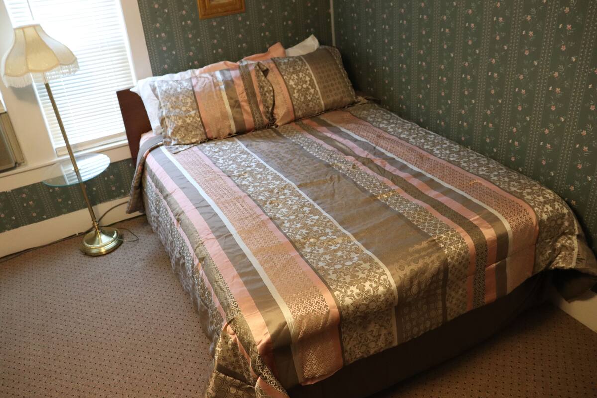 A neatly made bed with patterned bedding, a small round table with a lamp, and a chair.