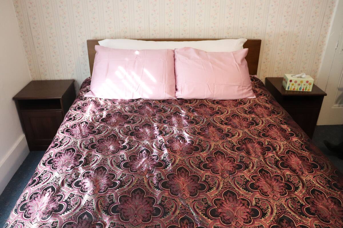 A bed with a patterned bedspread, two pillows, a nightstand with a tissue box, and a wall with floral wallpaper.