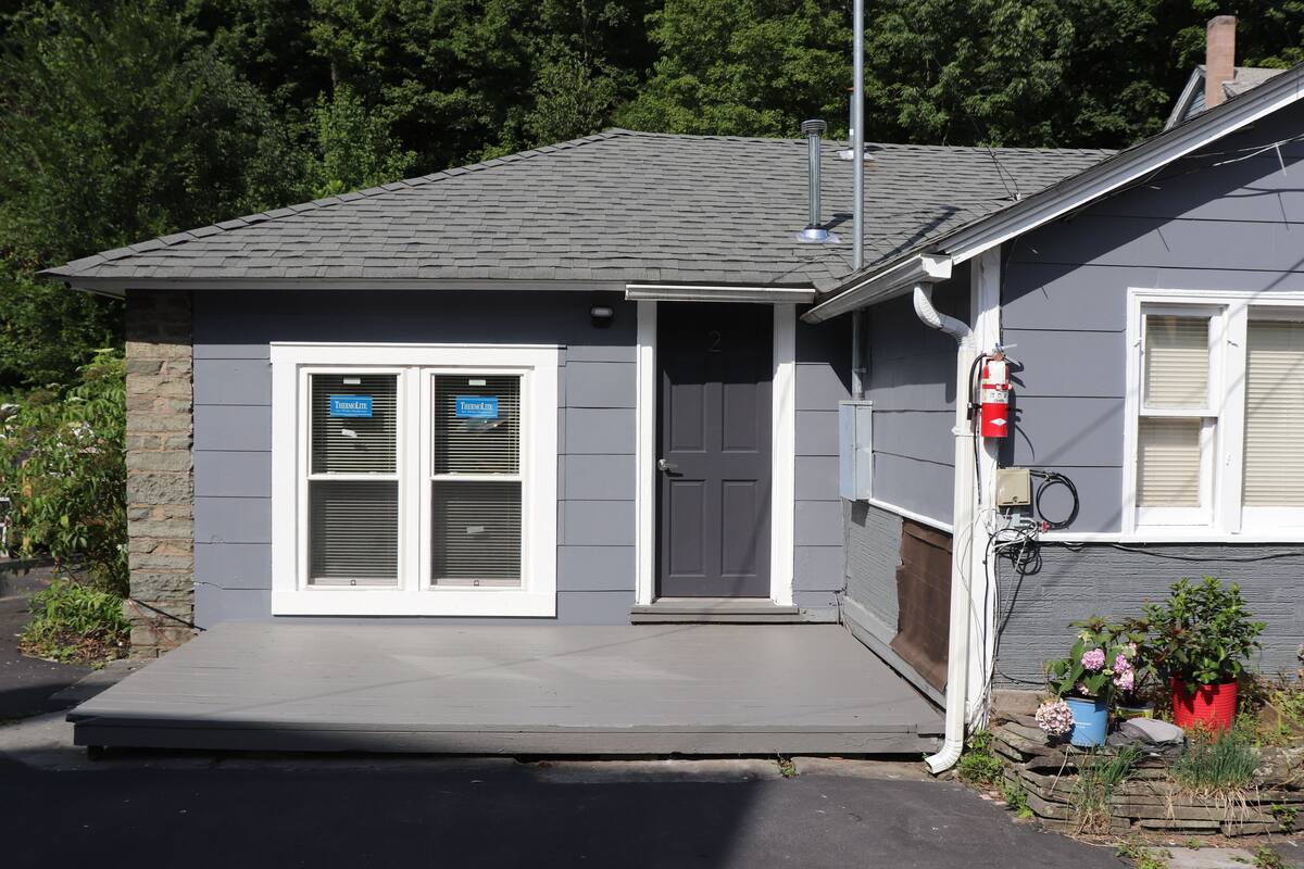 A small, single-story building with a gray exterior, a front door, and two windows with shutters.