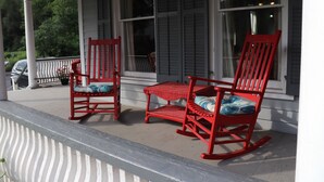 A porch with two rocking chairs and a small table, white railing, and a window with shutters.