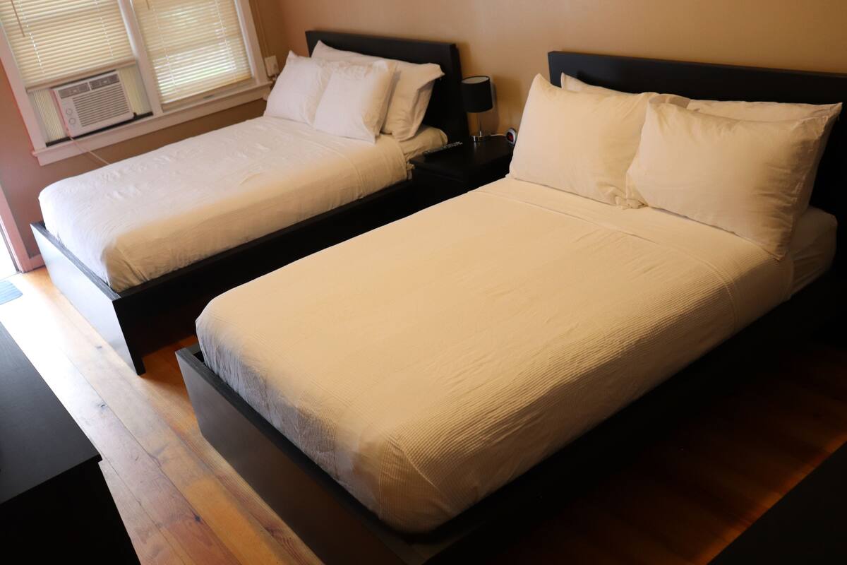 Two single beds with white linens, a nightstand with a lamp, and an air conditioning unit visible through the window.